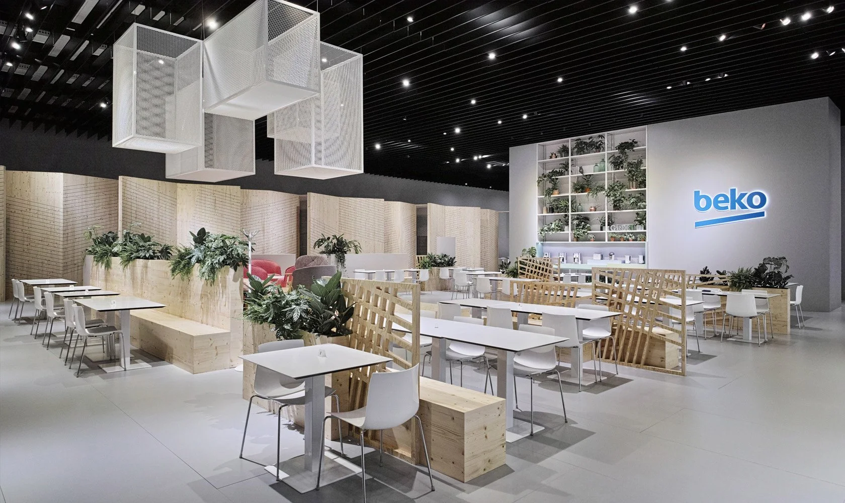 Modern retail store or cafe interior with white tables and chairs, wooden partition walls and decorative plants, illuminated by ceiling spotlights, with a large 'beko' logo on the wall.
