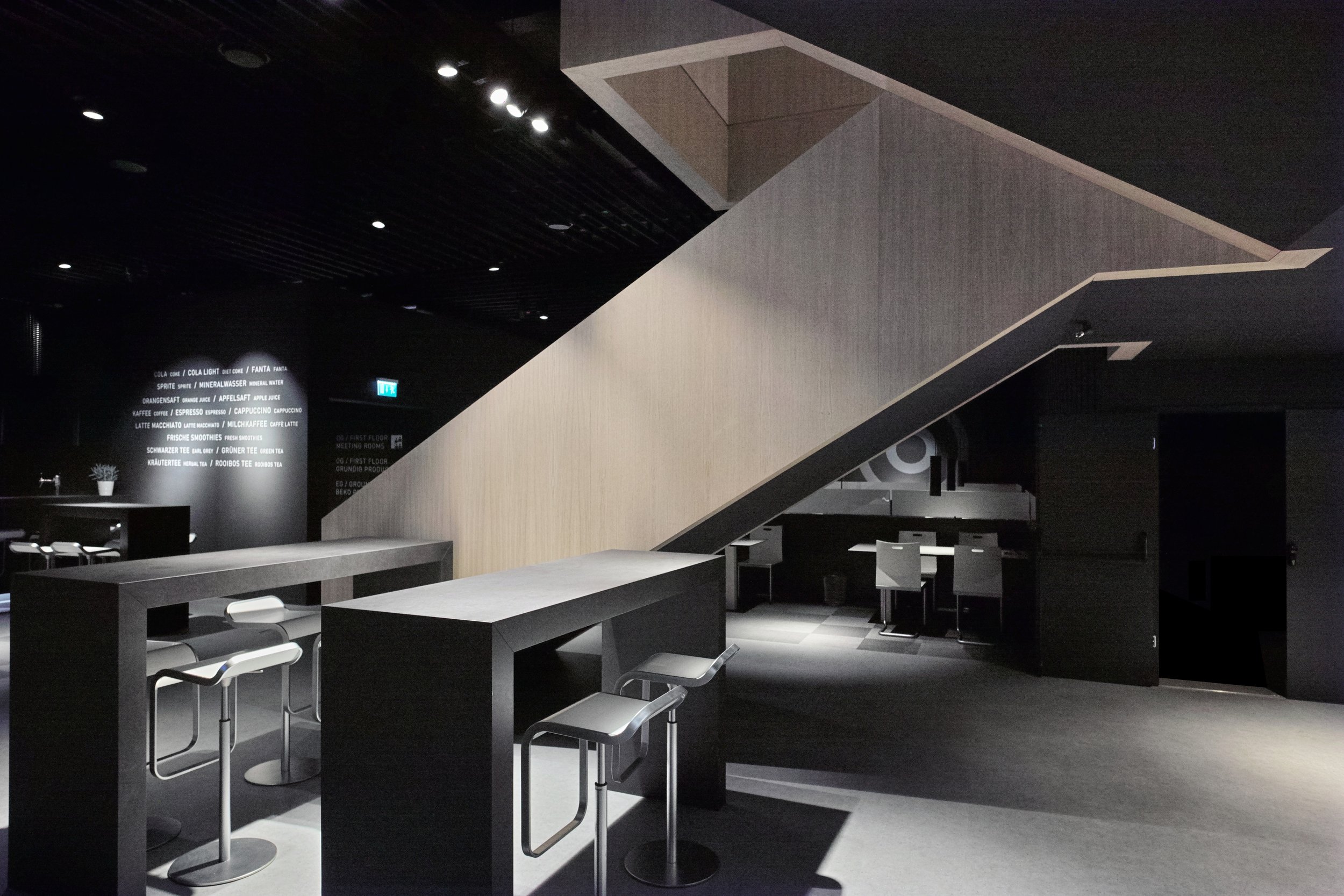 Modern interior of a cafe or lounge with high tables, bar stools, and a staircase leading to an upper level, with dark walls and minimal lighting.