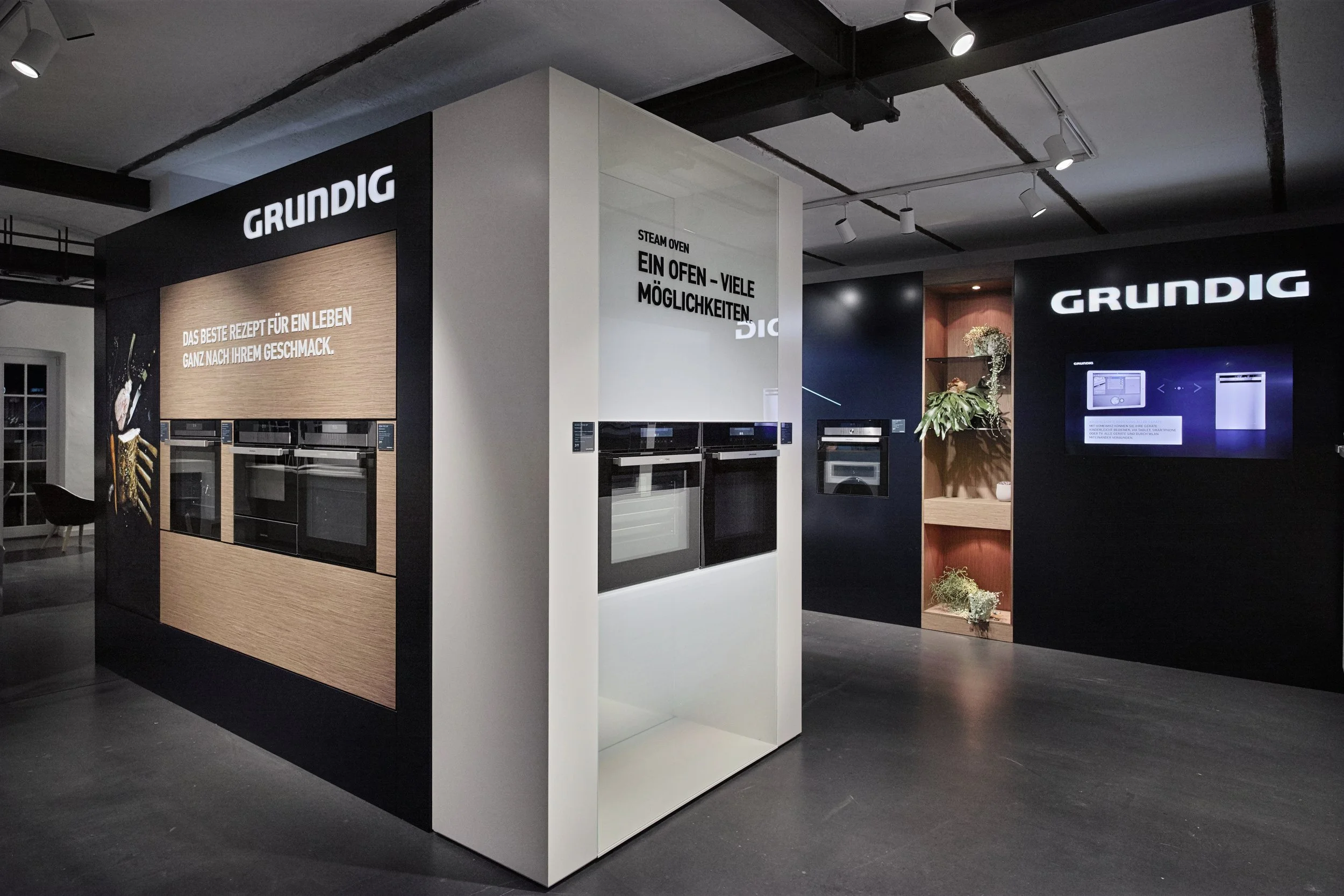 Display of Grundig kitchen appliances, including ovens, in a showroom with black and white walls and decorative plants.