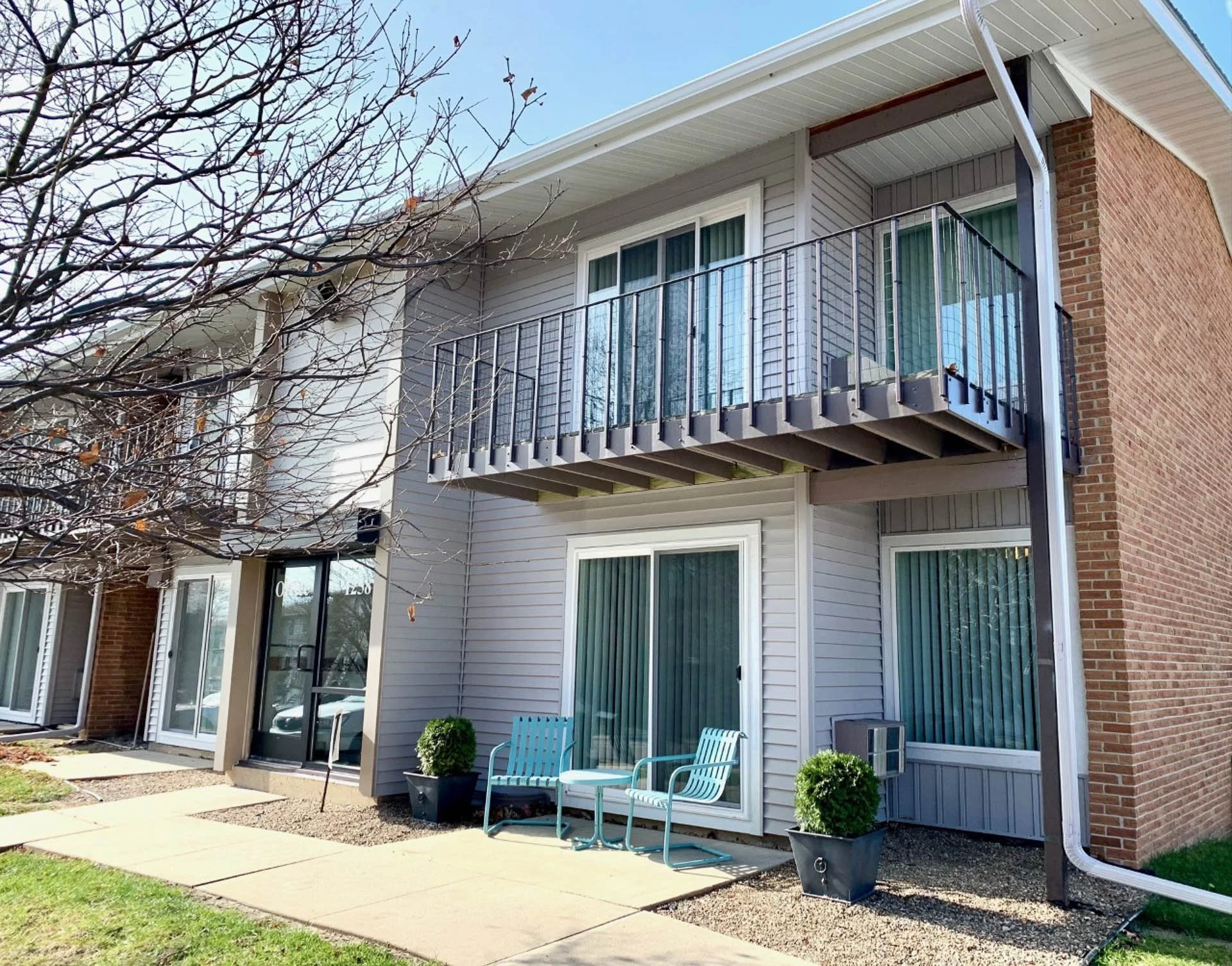Exterior of a two-story residential building with a balcony, glass sliding doors, and patio furniture outside on a paved walkway, with a leafless tree in the foreground.