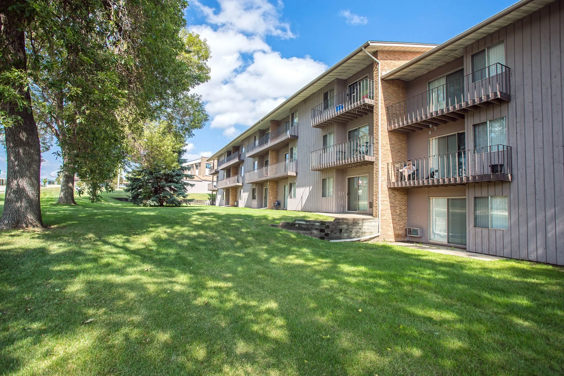 Apartment building with balconies surrounded by green lawn and trees under a blue sky with clouds.