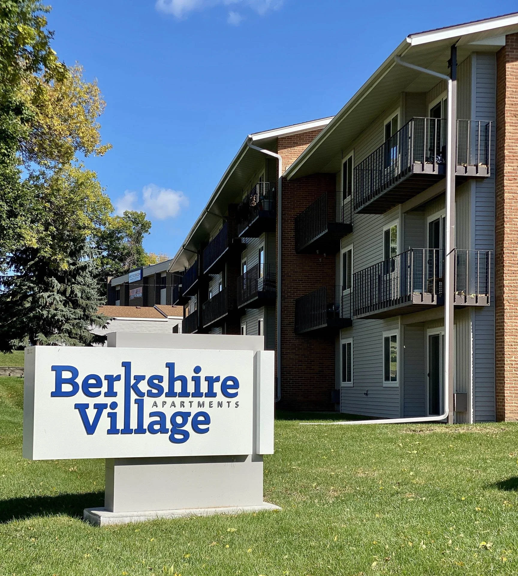 Apartment complex with a sign that reads Berkshire Village Apartments, surrounded by green grass and trees, with balconies on the buildings and a sunny sky in the background.