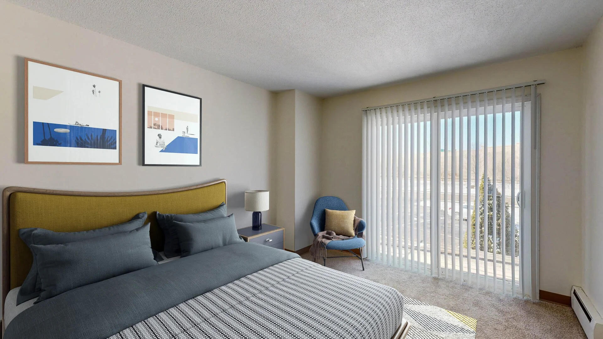 A bedroom with a bed that has a yellow headboard, dark gray bedding, and gray pillows. There is a nightstand with a lamp next to the bed. A blue armchair with a yellow pillow and a blanket is near the window with vertical blinds. Two framed abstract 