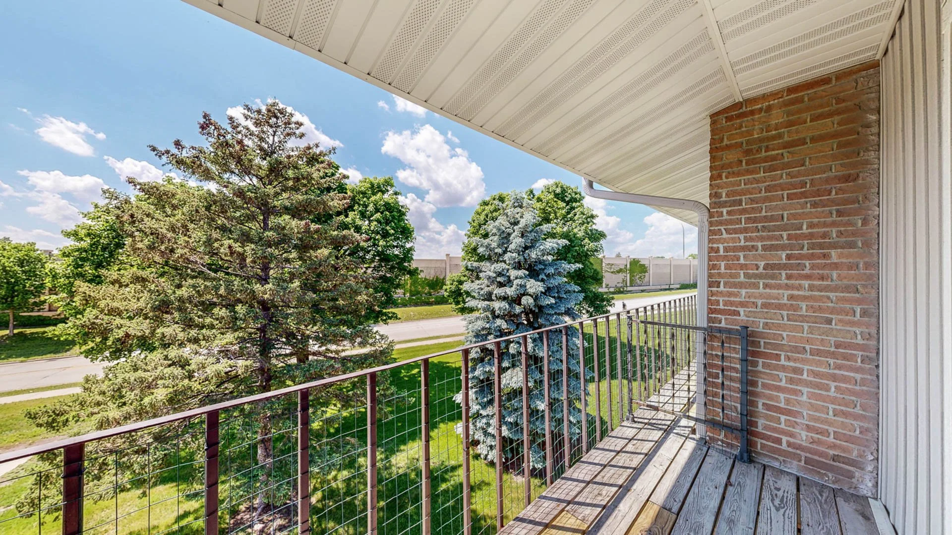 View from a balcony showing green trees, blue sky with white clouds, a road, a brick wall, and a metal railing.
