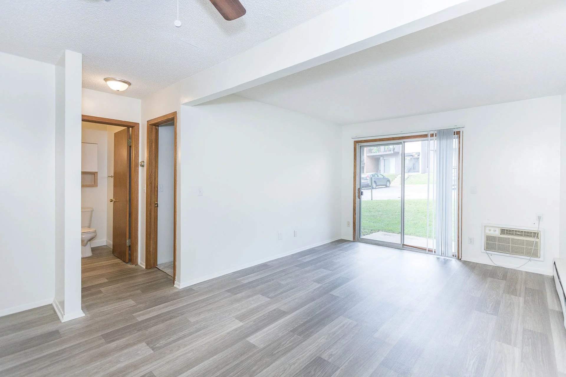 Empty living room with sliding glass door leading outside, wood flooring, and a small wall heater.