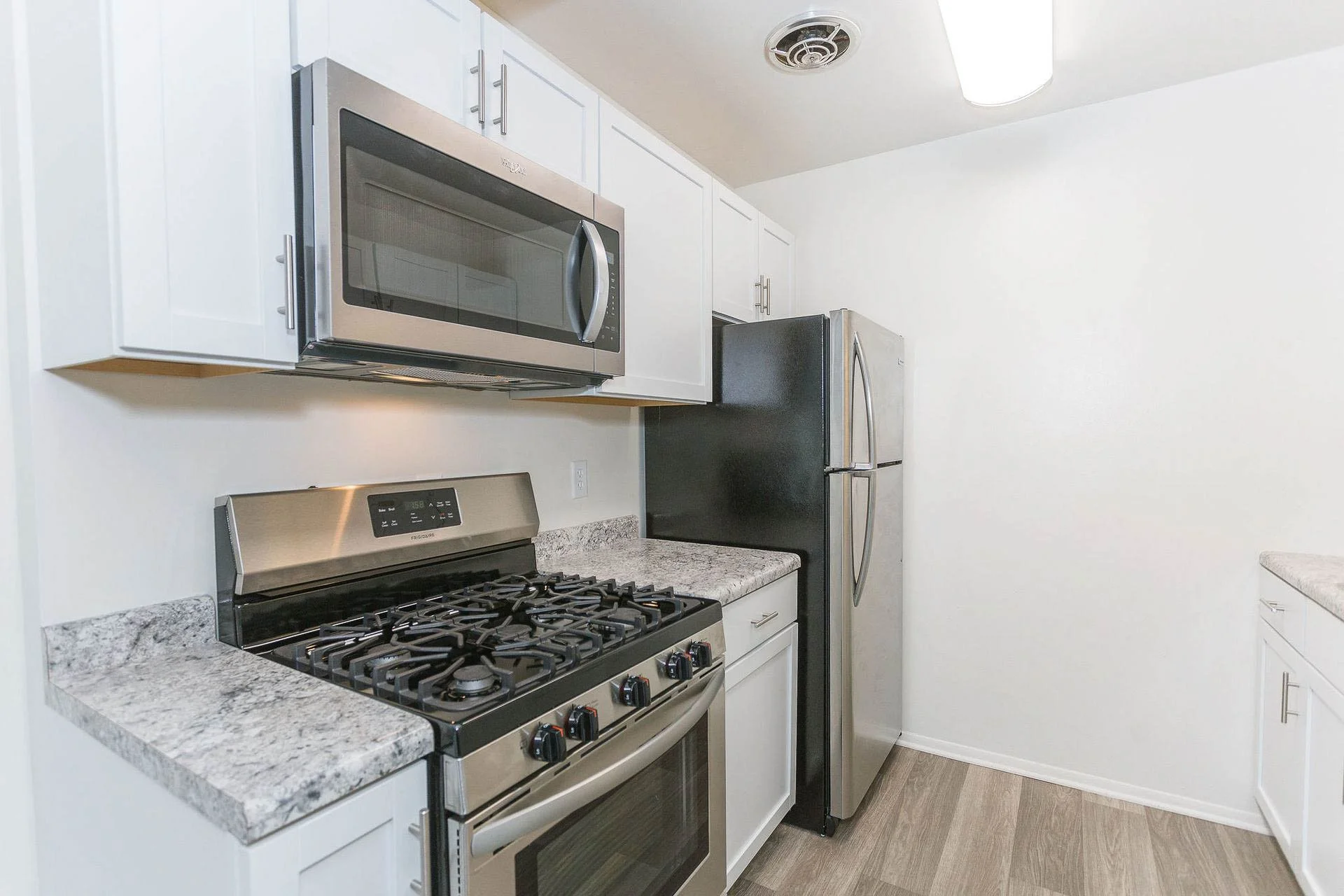 Kitchen with white cabinets, granite countertops, stainless steel microwave above a gas stove, with a refrigerator to the right, on a wood floor.