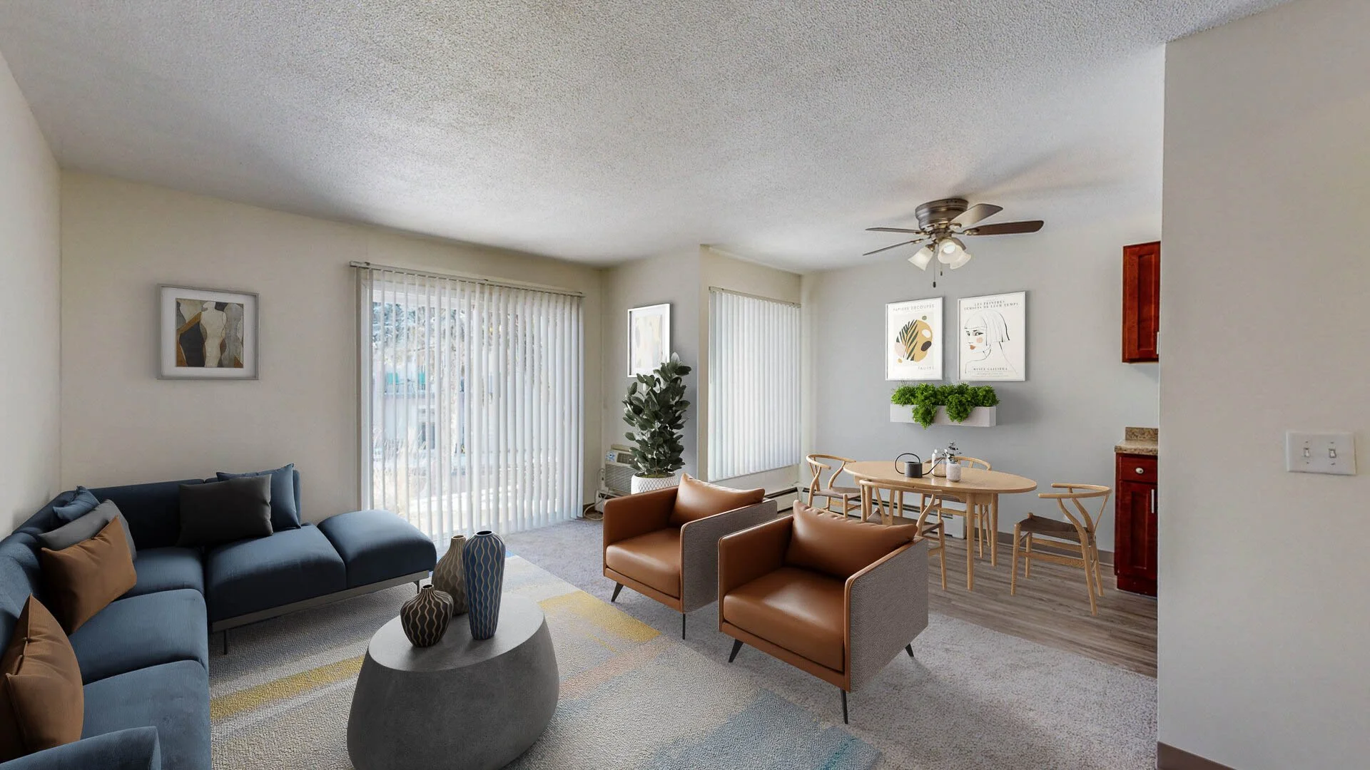 Living room with blue sofa, tan and brown armchairs, a coffee table with vases, a dining table with chairs, and a ceiling fan, illuminated by natural light through vertical blinds.