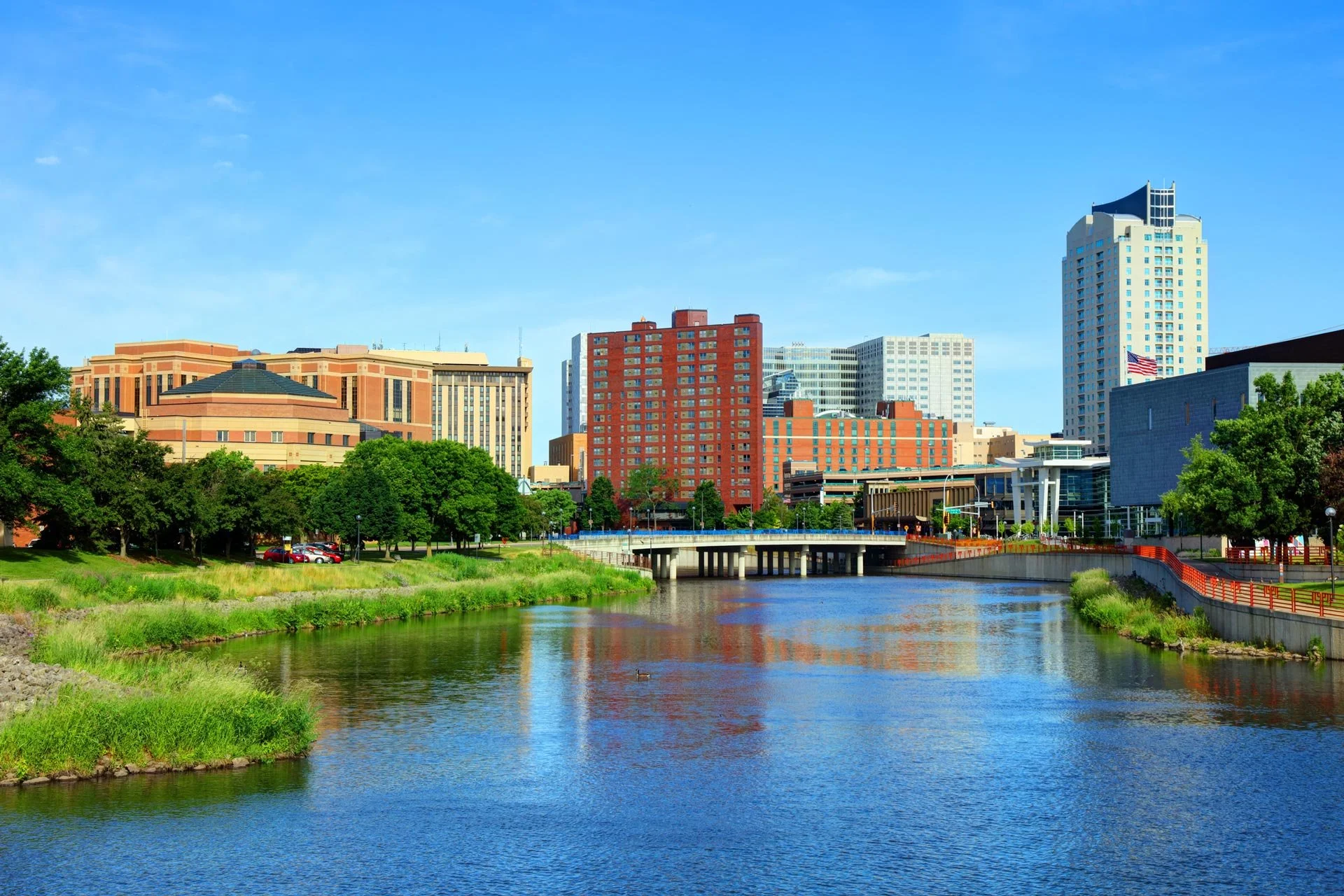 Rochester city skyline with tall buildings, a river in the foreground, and a clear blue sky.