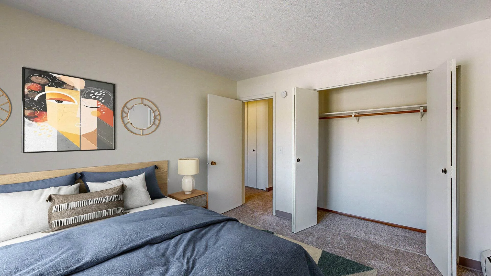 A bedroom with a neatly made bed featuring multiple pillows, a small wooden nightstand with a lamp, abstract art hanging on the wall, and an open closet with sliding doors.