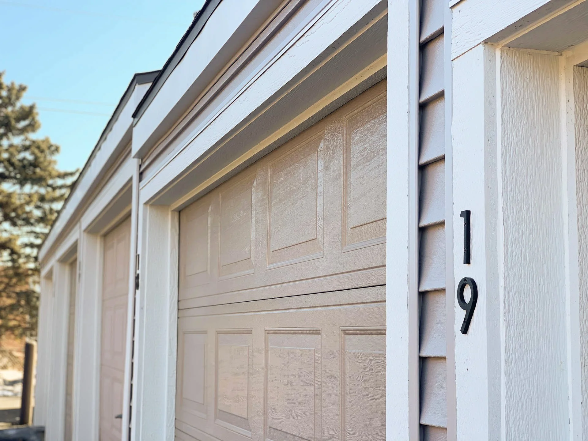 Close-up of a beige garage door and white house exterior, with house number 19 on the side and some trees in the background.