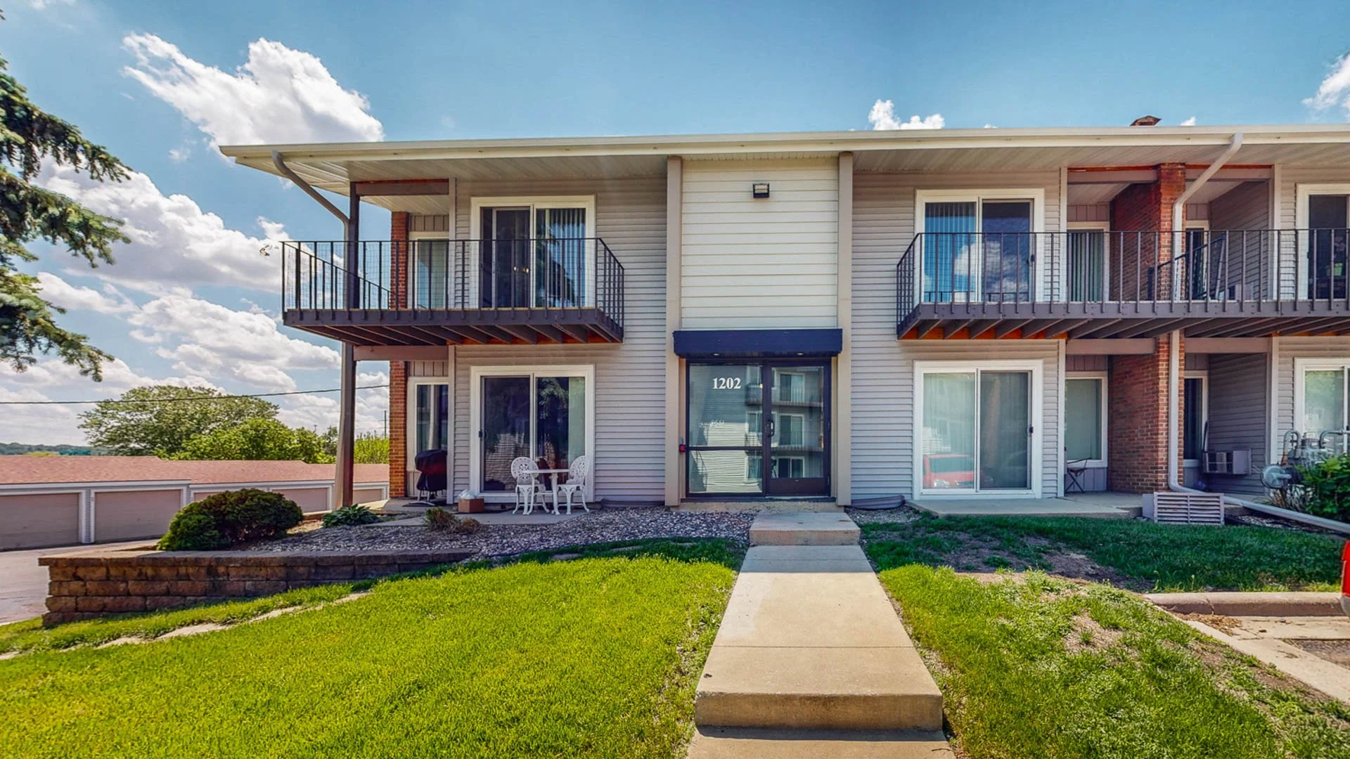 Exterior of a two-story apartment building with balconies, white siding, sliding glass doors, and a green lawn with a concrete walkway leading to the front door, under a blue sky with clouds.