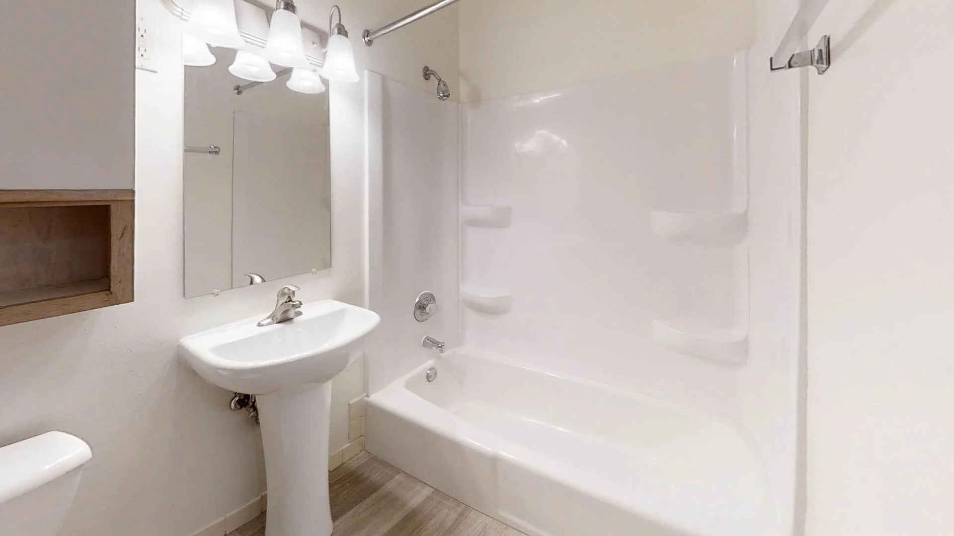 A small bathroom with a white bathtub, showerhead, and built-in shelves, a pedestal sink with a mirror and light fixtures above, and a partial view of a toilet with wood flooring.