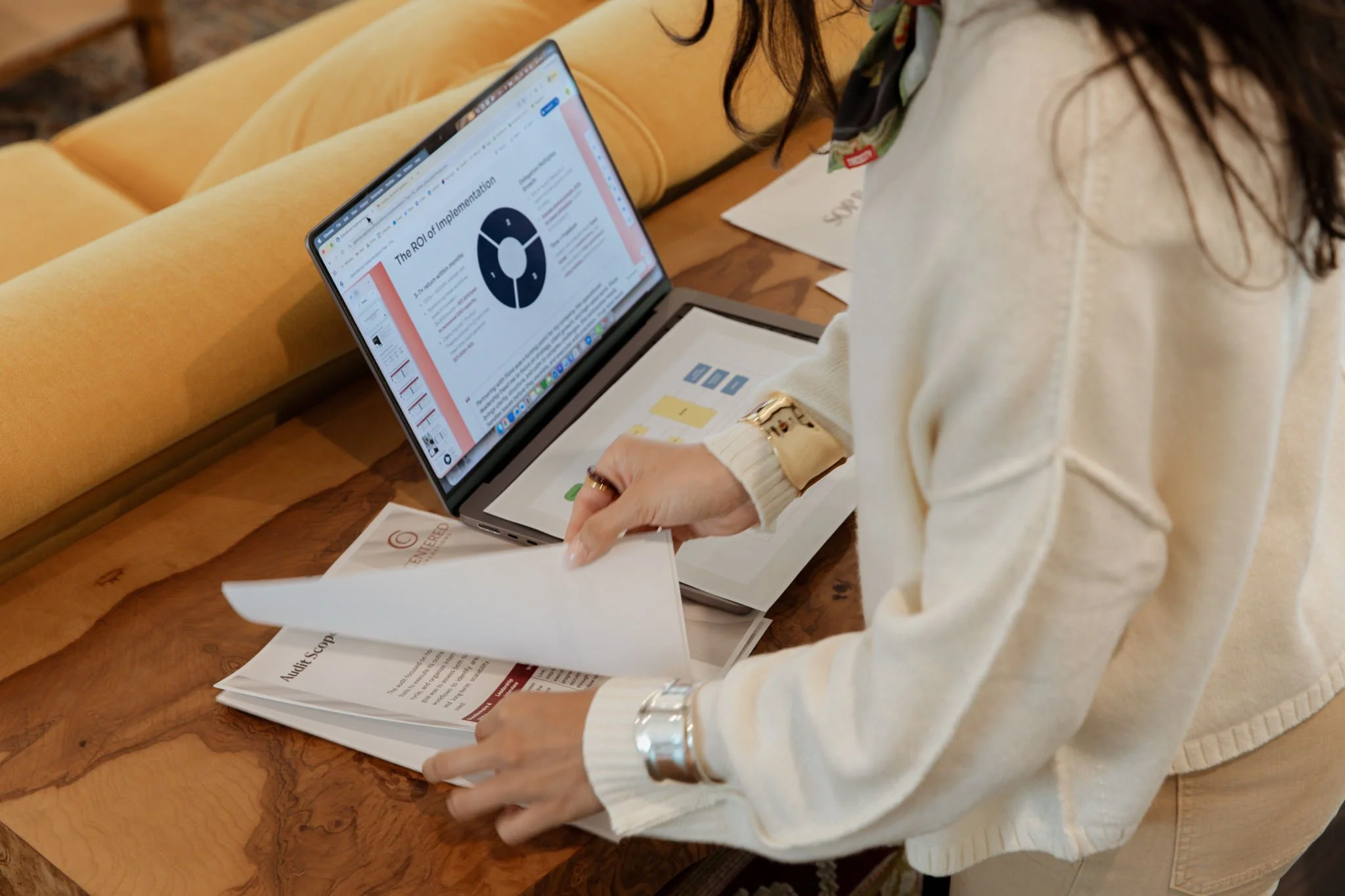 A woman wearing a white sweater and a silk scarf looks at documents and a laptop screen displaying a presentation on ROI, sitting at a wooden table with papers and folders.
