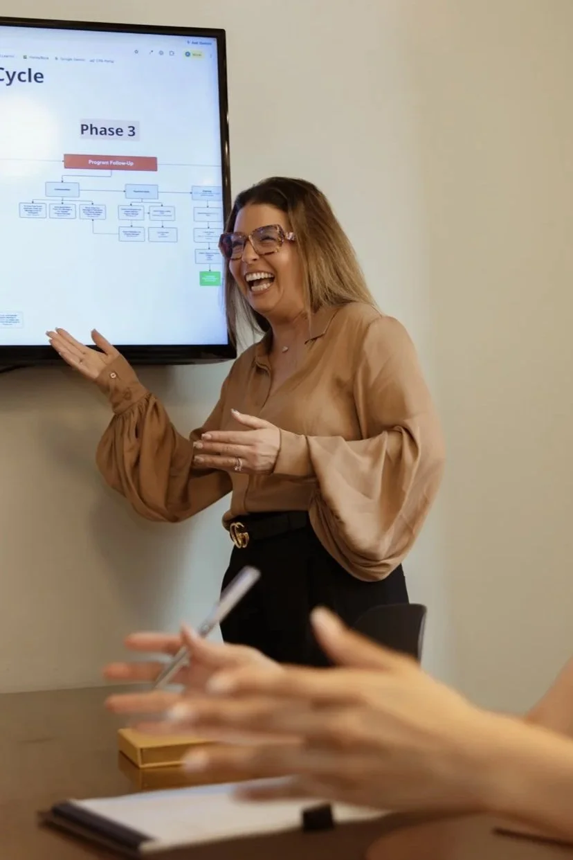 A woman with shoulder-length hair and glasses, wearing a beige blouse, is smiling and pointing at a presentation on a large screen in a meeting room.