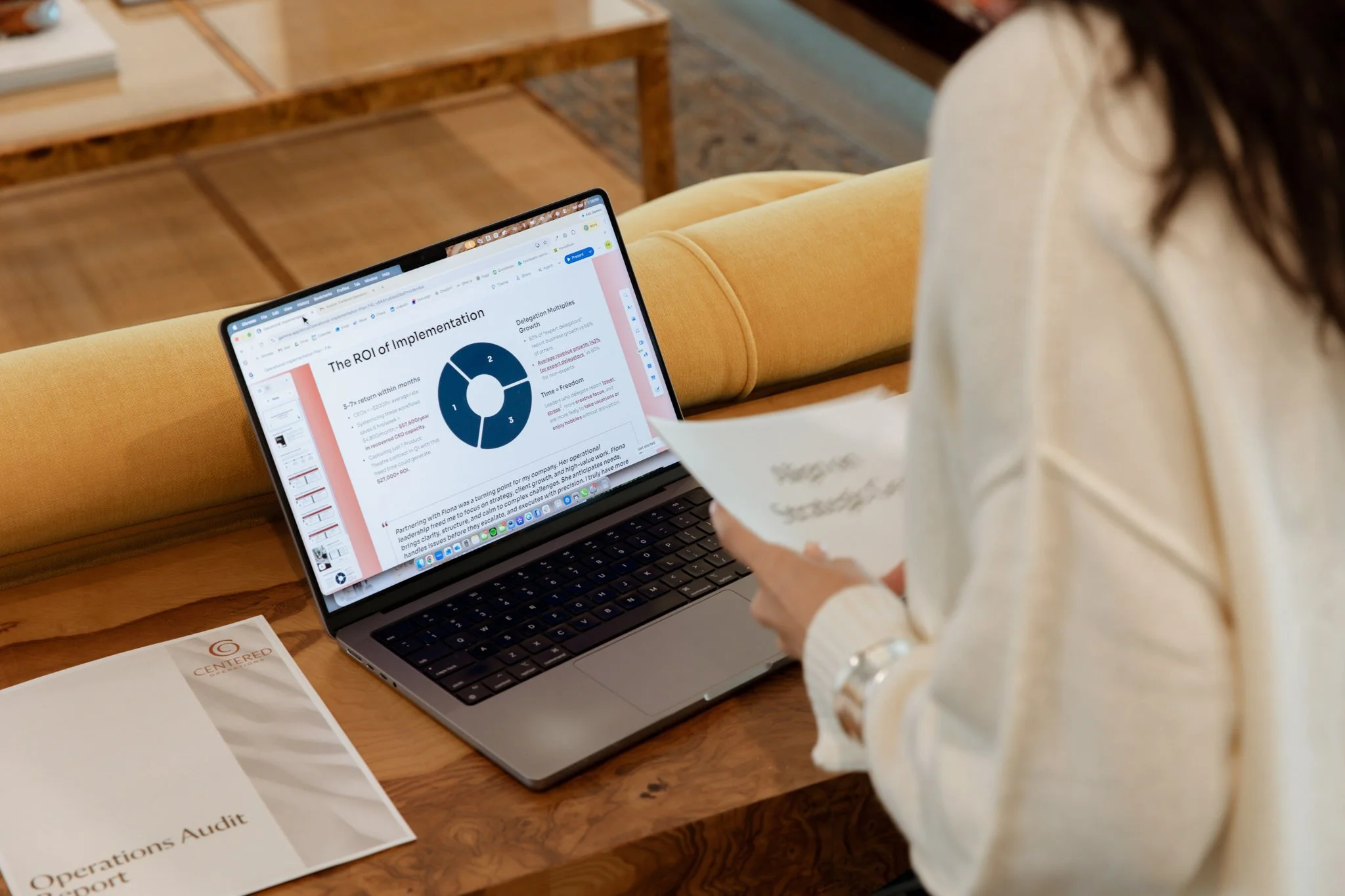 A person in a white shirt using a laptop on a wooden table, with a presentation slide titled 'The ROI of Implementation' visible on the screen.
