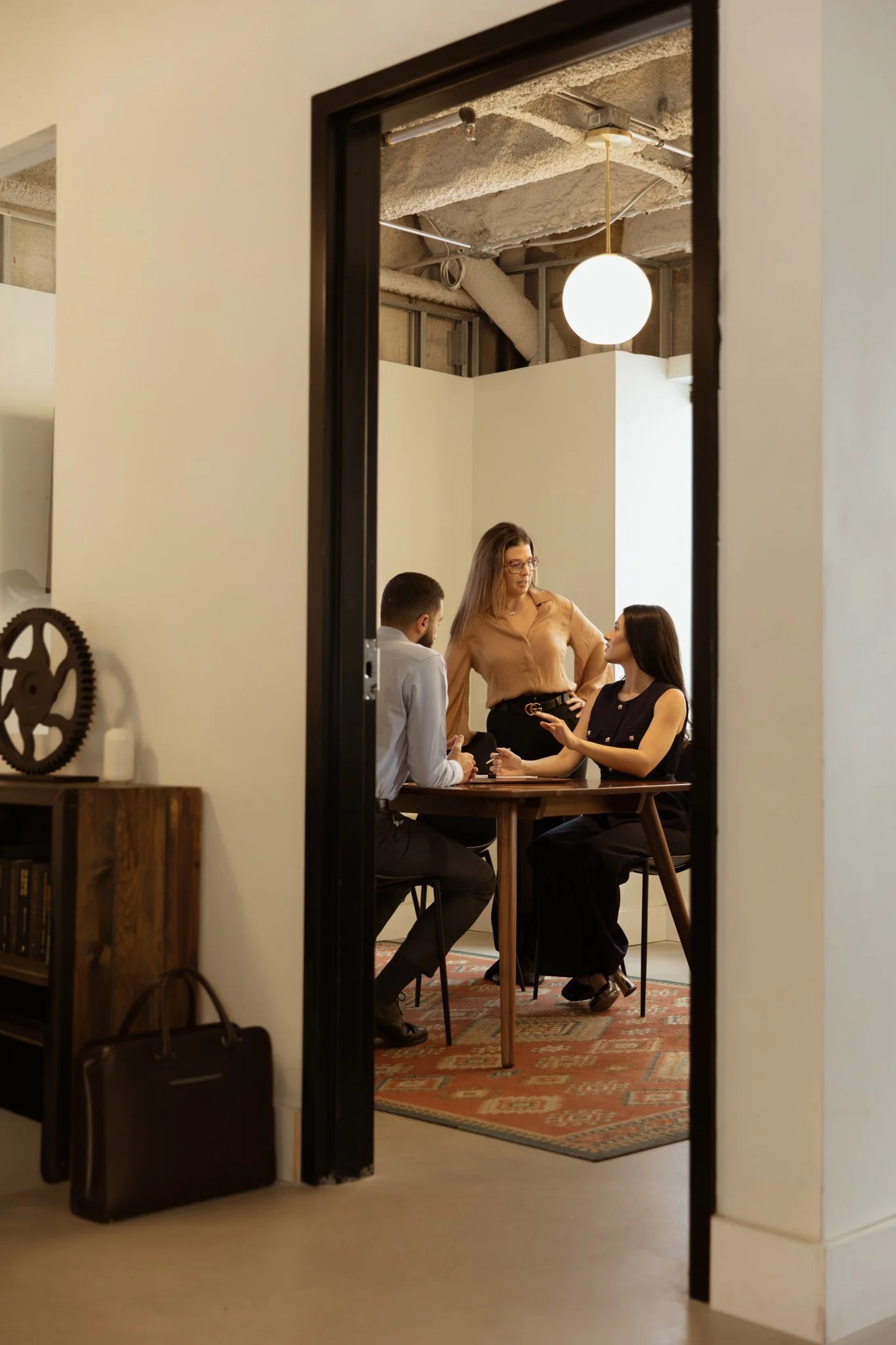 A group of three people inside a room, viewed through a doorway. Two women and a man and the group appear to be engaged in a conversation around a table. The room has an industrial ceiling with exposed beams and a white spherical hanging light. The setting appears professional, possibly a meeting or discussion.