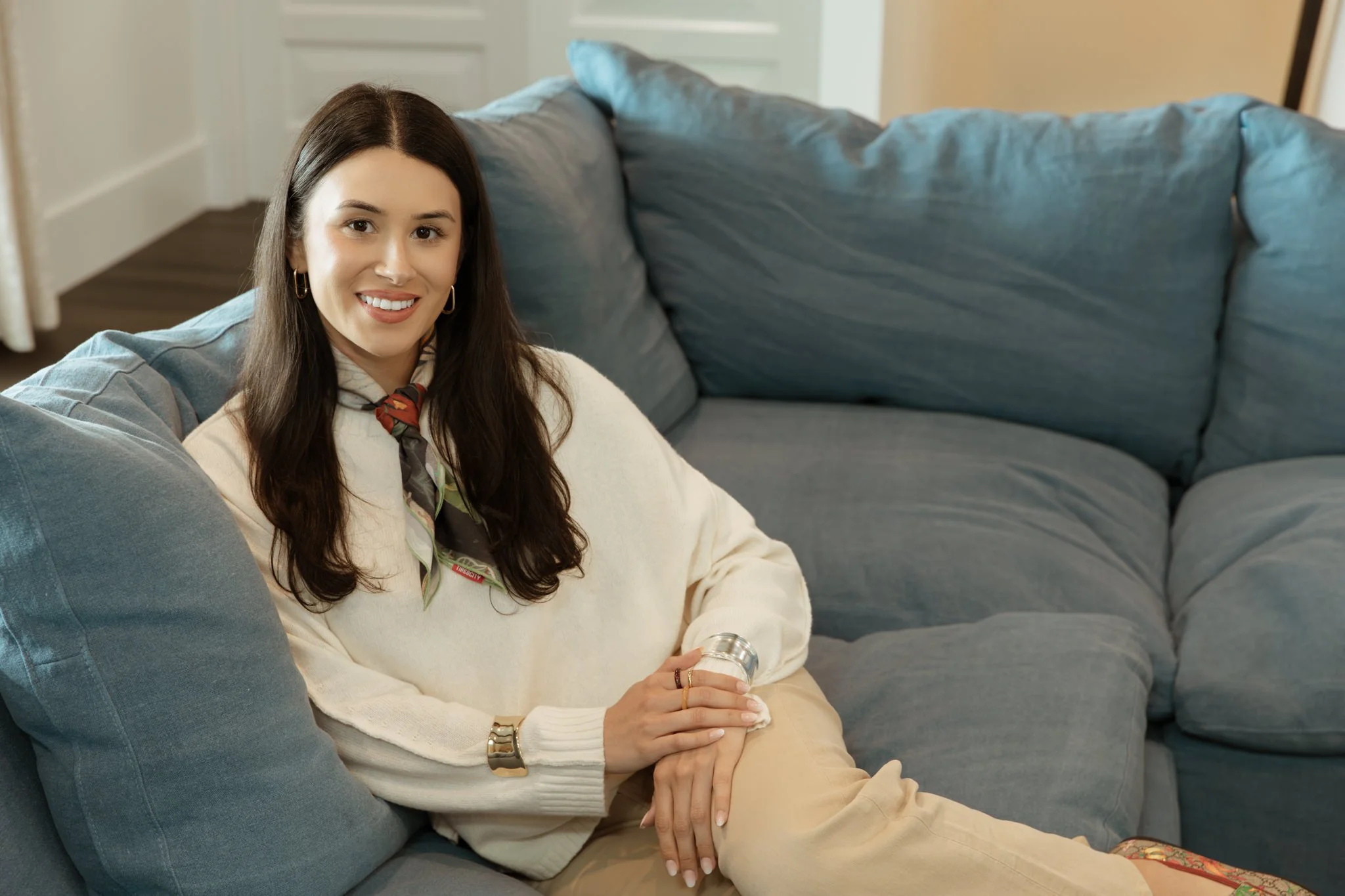 A young woman with long dark hair, wearing a white sweater, seated on a blue couch with large pillows, smiling and holding a small jar, in a room with a door and beige walls.