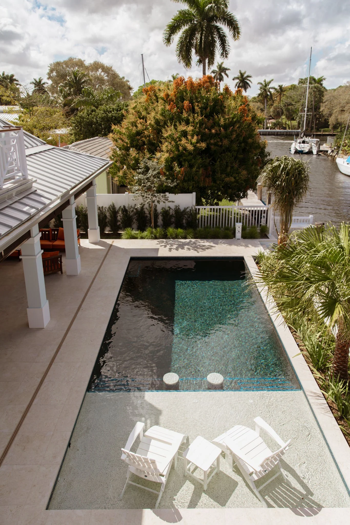 A backyard with a rectangular pool, a white patio table with six chairs, lush green palm trees, a white fence, and a canal with boats docked on the water under a partly cloudy sky.