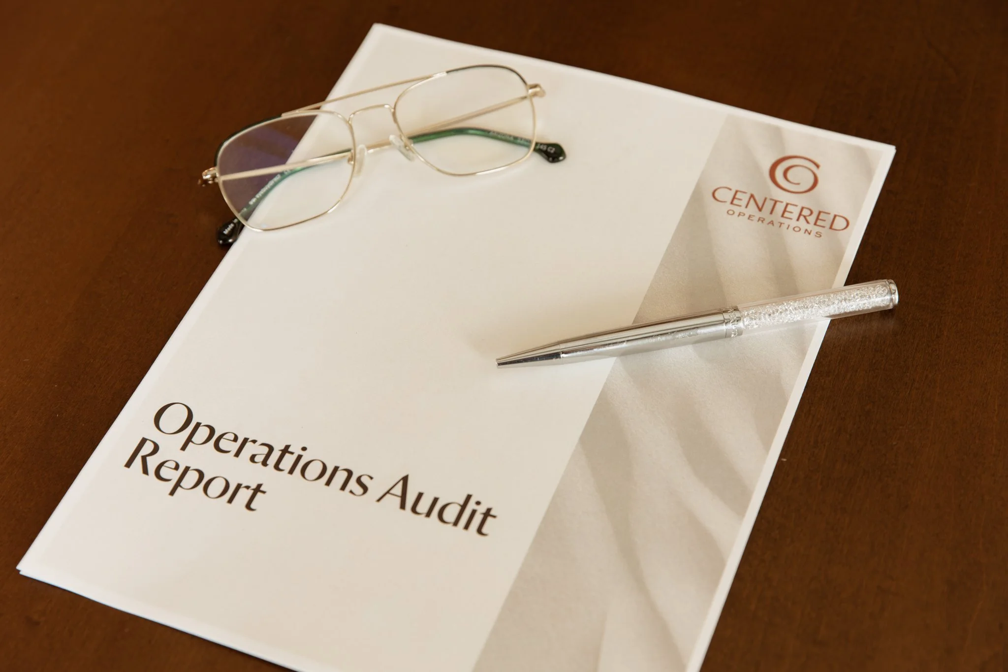 A pair of eyeglasses with a thin metal frame, a metallic pen with a clear, textured body, resting on a white report titled 'Operations Audit Report' on a wooden surface, with a paper behind the report displaying the logo and name 'Centered Operations'.