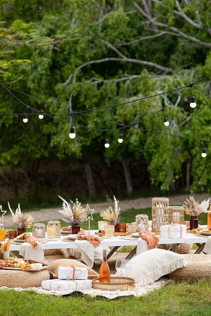 Outdoor picnic table decorated with flowers, candles, and gift boxes, surrounded by cushions on the grass, with string lights overhead and trees in the background.