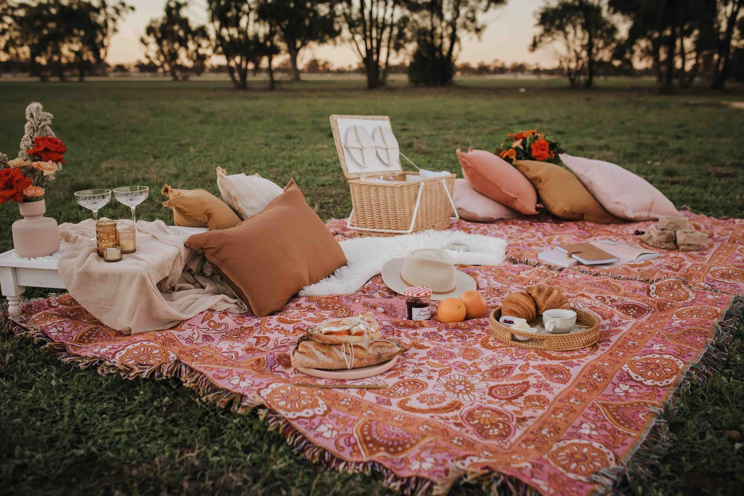 A picnic setup on a patterned blanket in a grassy field at sunset with cushions, flowers, glasses, a wicker basket, pastries, fruits, and a hat.