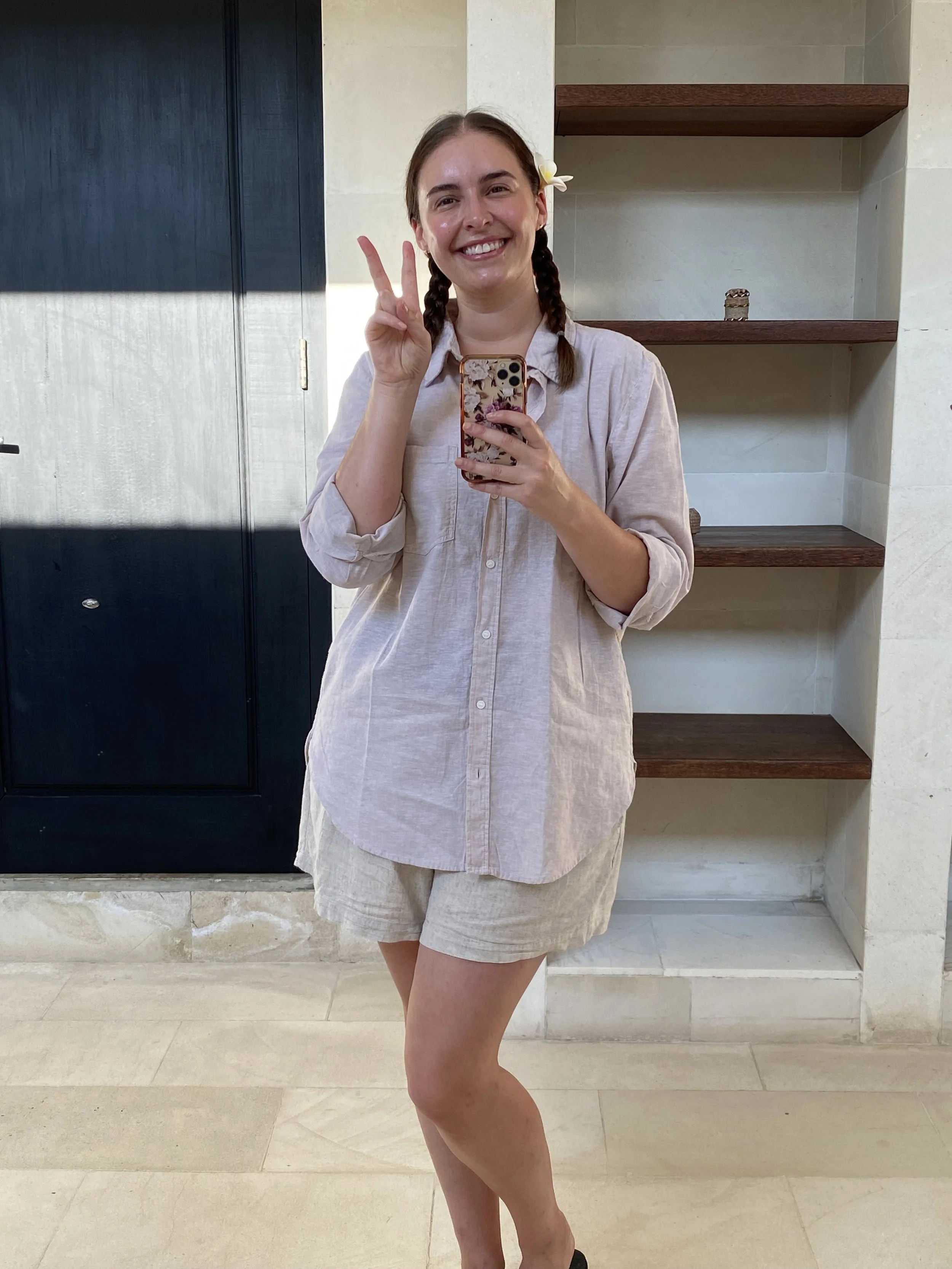 Young woman with braided hair, wearing a light-colored shirt and shorts, smiling and making a peace sign while taking a selfie indoors.