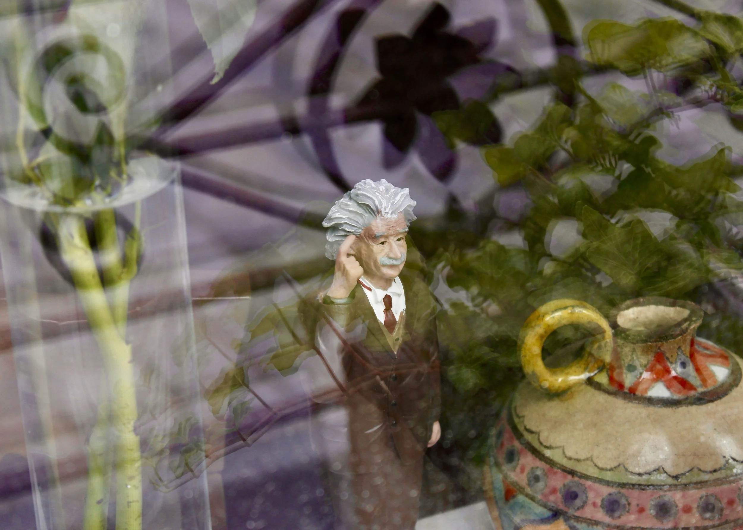A small figurine of Albert Einstein with white hair, mustache, and a suit, standing among plants and decorative pottery, seen through a glass surface with reflections.