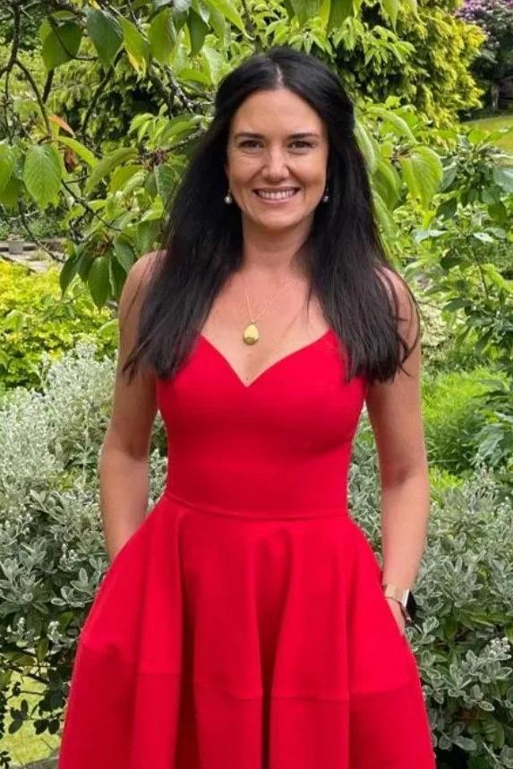 Woman in a red dress standing outdoors among green trees and lush foliage.