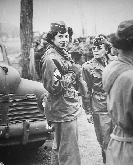 Two women soldiers in military uniforms talking outdoors during wartime, with a military vehicle in the background.