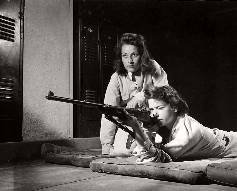 Two women in a locker room, with one lying on her stomach aiming a rifle and the other standing behind her, observing.
