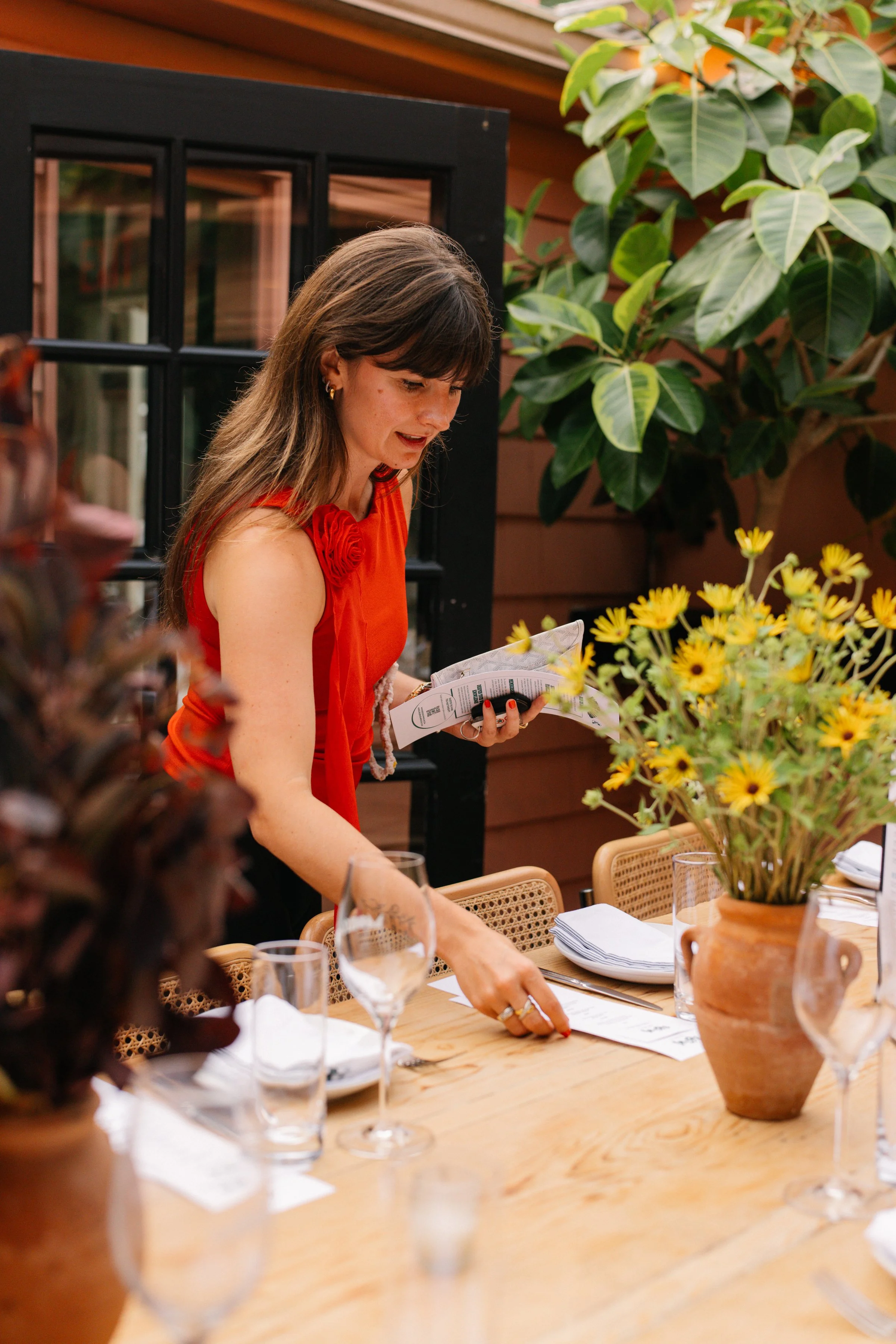 A woman in an orange sleeveless top with a rose decoration is standing at a dining table, reading a menu. The table has a large vase with yellow flowers, wine glasses, plates, and napkins. The setting appears to be indoors with plants and wooden walls in the background.