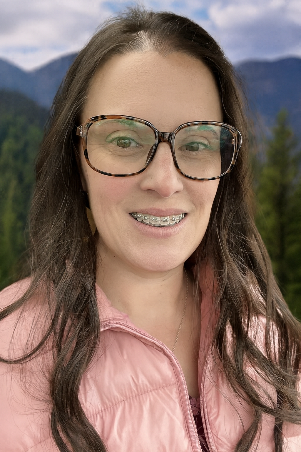 A woman with long brown hair wearing large tortoiseshell glasses, braces, and a pink jacket, smiling in front of a mountain landscape with trees and mountains in the background.