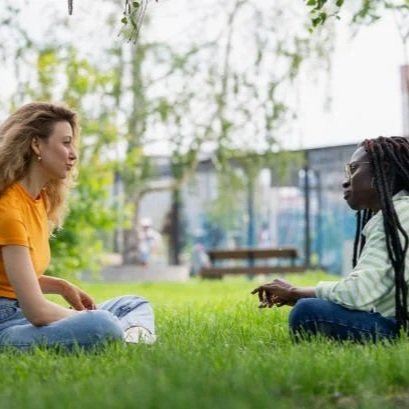 Two women sitting on grass and talking in a park with trees and a bench in the background.