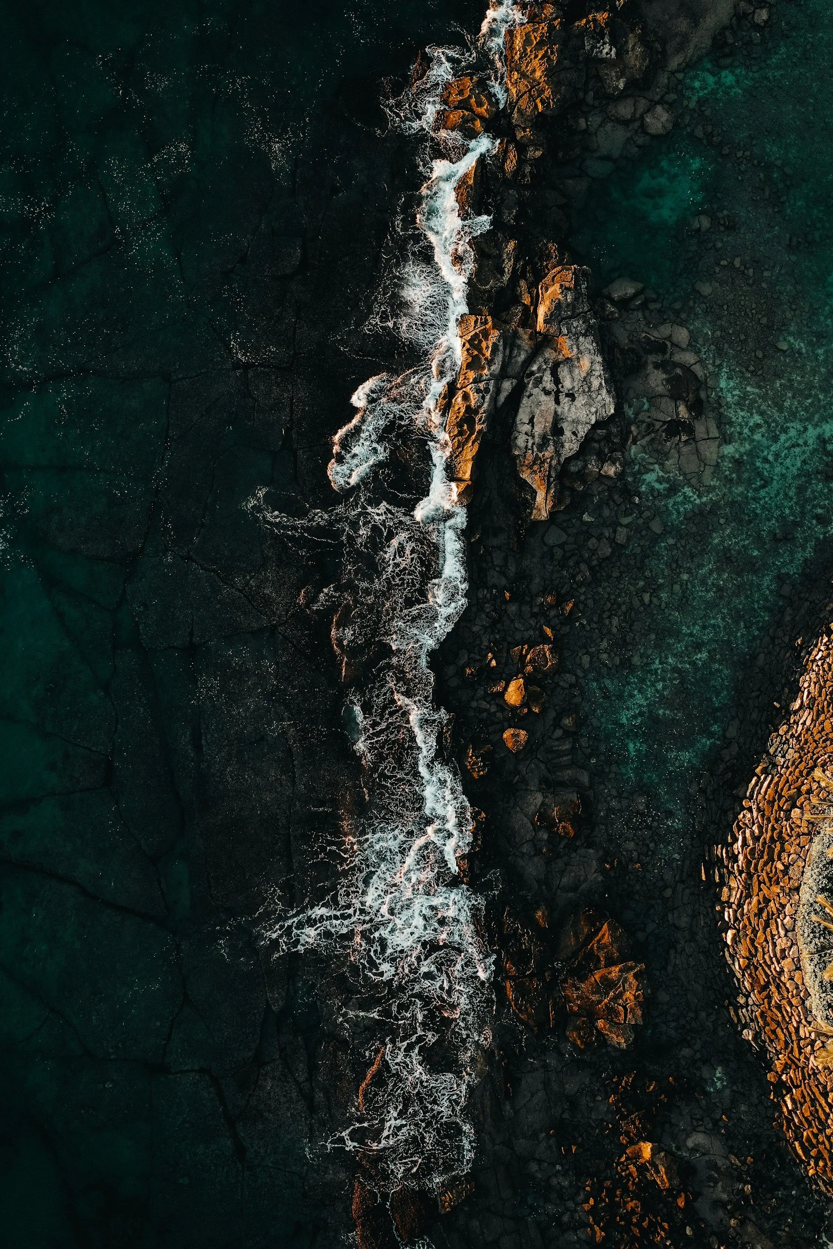 An aerial view of rocky coastline with waves crashing against rocks and dark ocean water nearby.