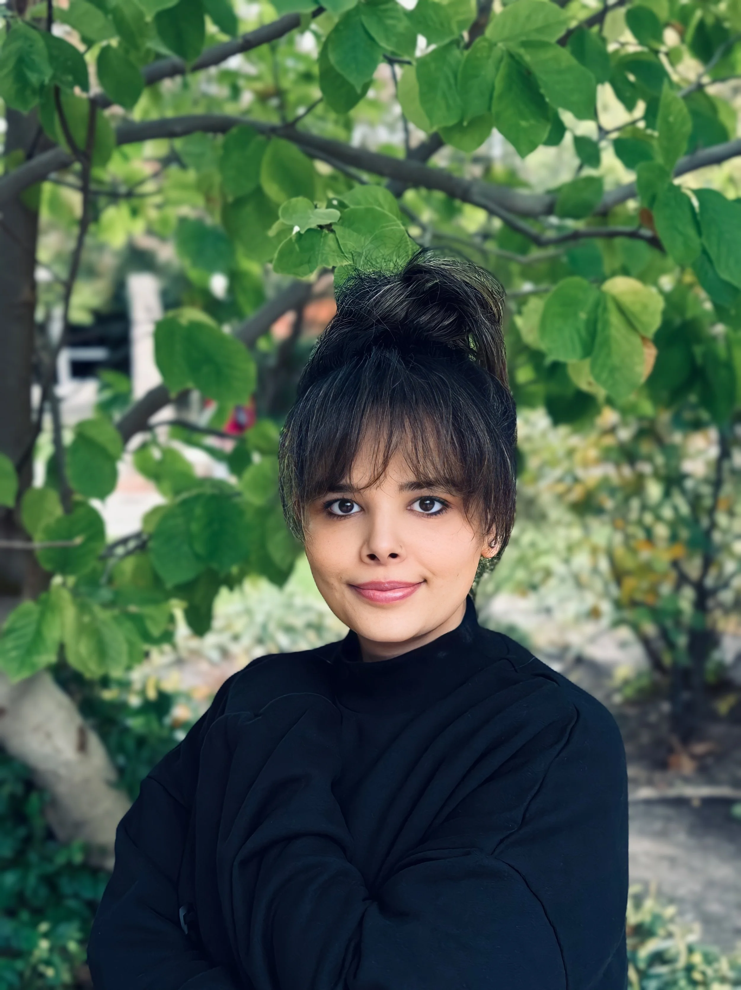 A young woman with dark hair styled in a high bun, wearing a black top, standing outdoors in front of green foliage and trees.