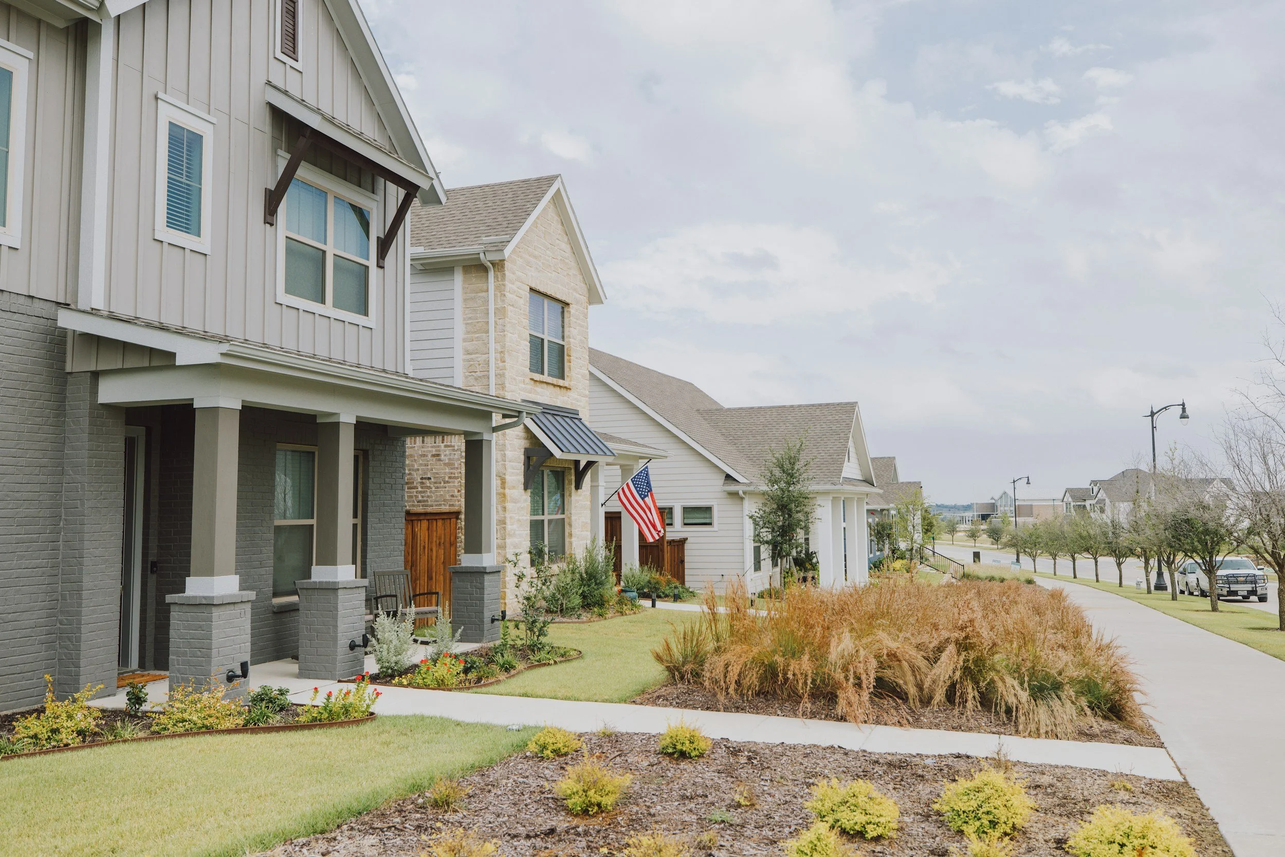 A row of modern suburban houses with well-maintained lawns and landscaped gardens, American flags in front yards, and a sidewalk lined with young trees, under a partly cloudy sky.
