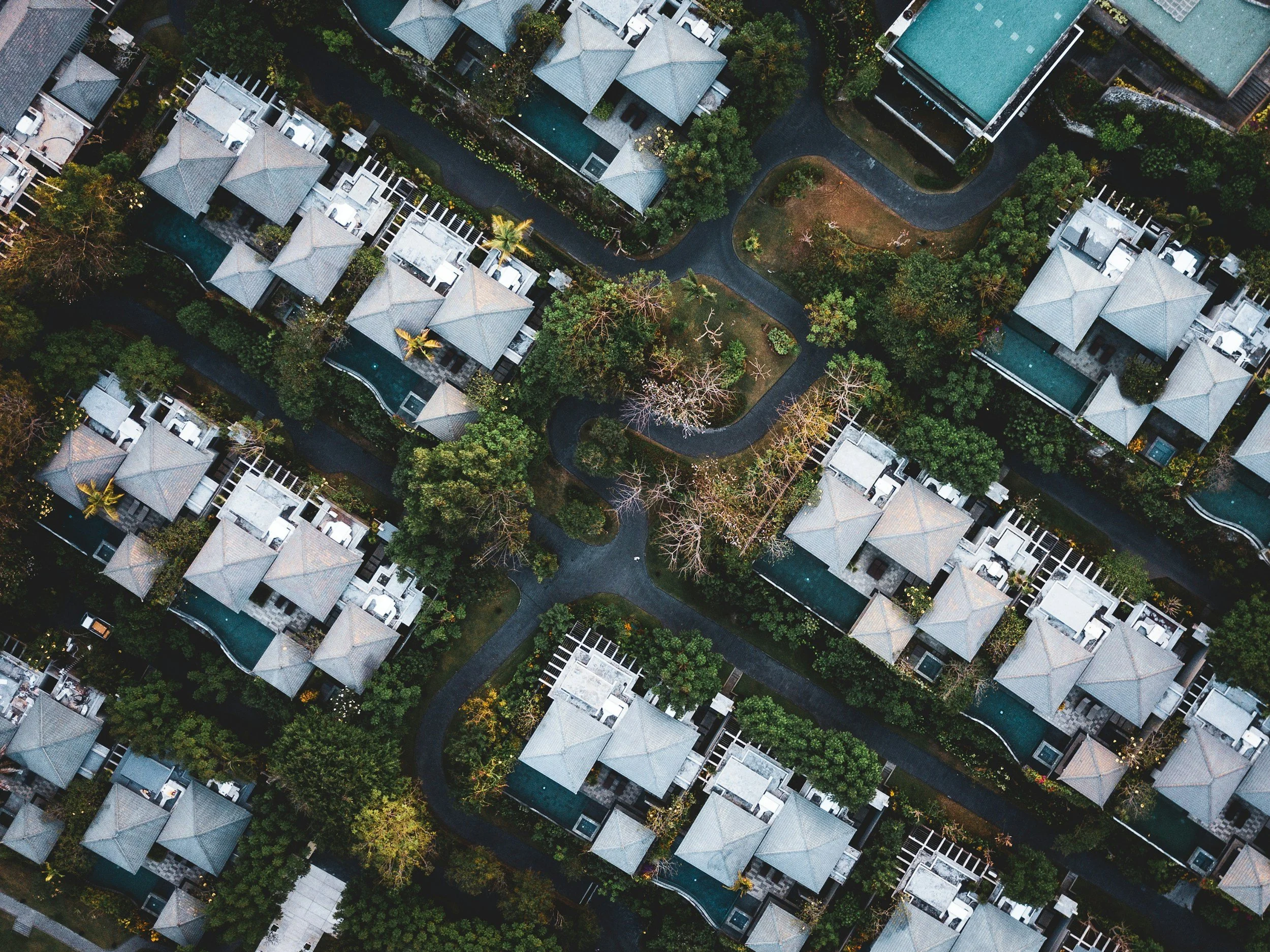 An aerial view of a residential neighborhood with multiple houses featuring gray roofs, surrounded by green trees and winding roads.