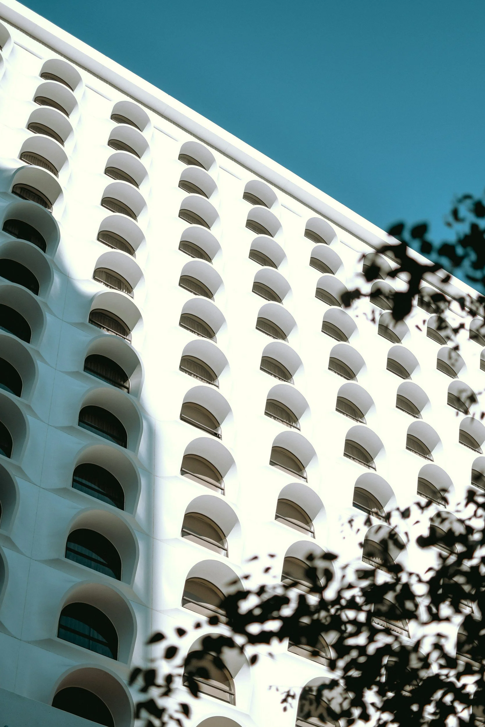 A tall white building with multiple rounded balconies on each floor, viewed from below against a clear blue sky. Some black leaves are visible in the foreground.