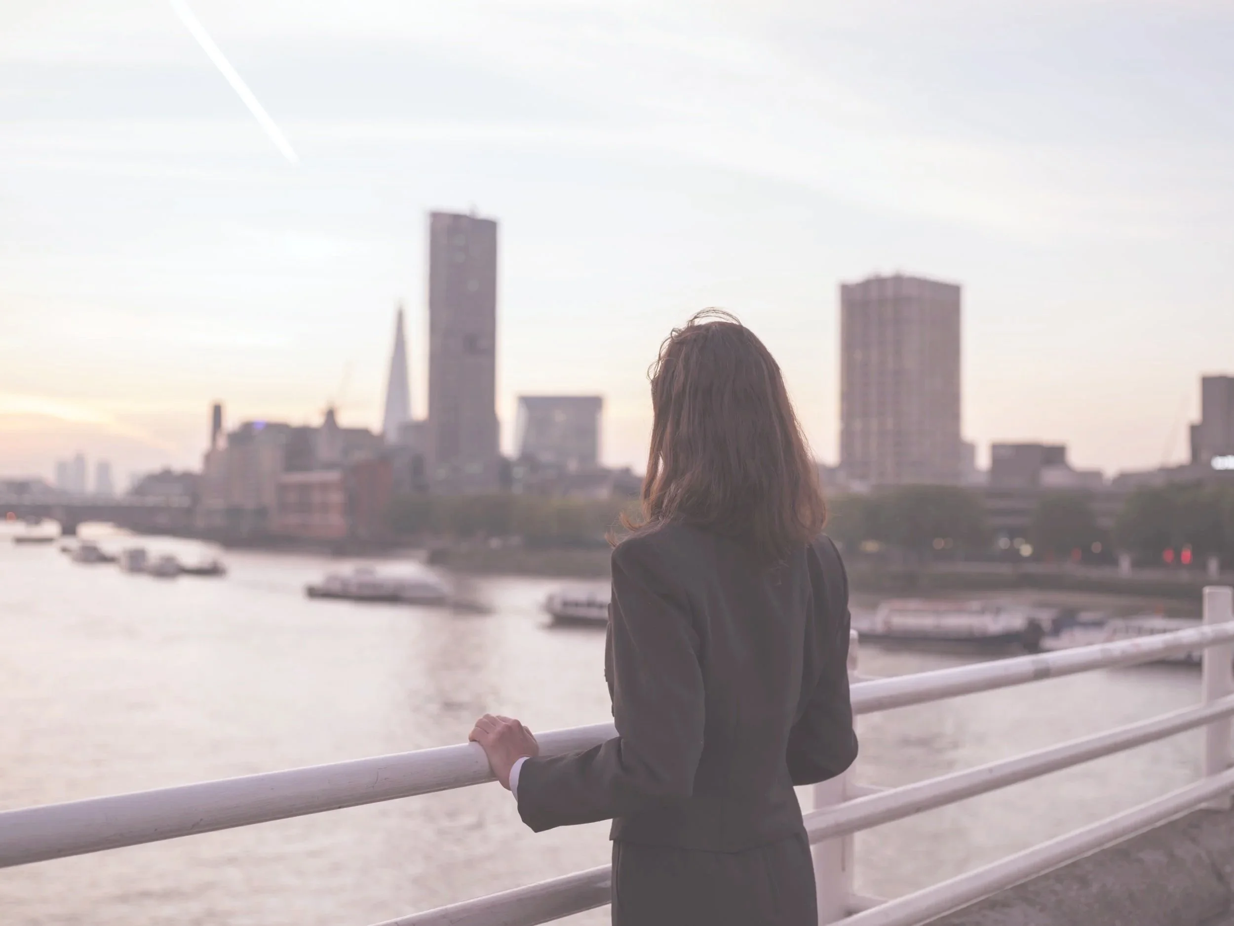 A woman with shoulder-length dark hair, dressed in a black blazer, stands on a riverside walkway, looking at the city skyline with tall buildings during sunset or early evening.