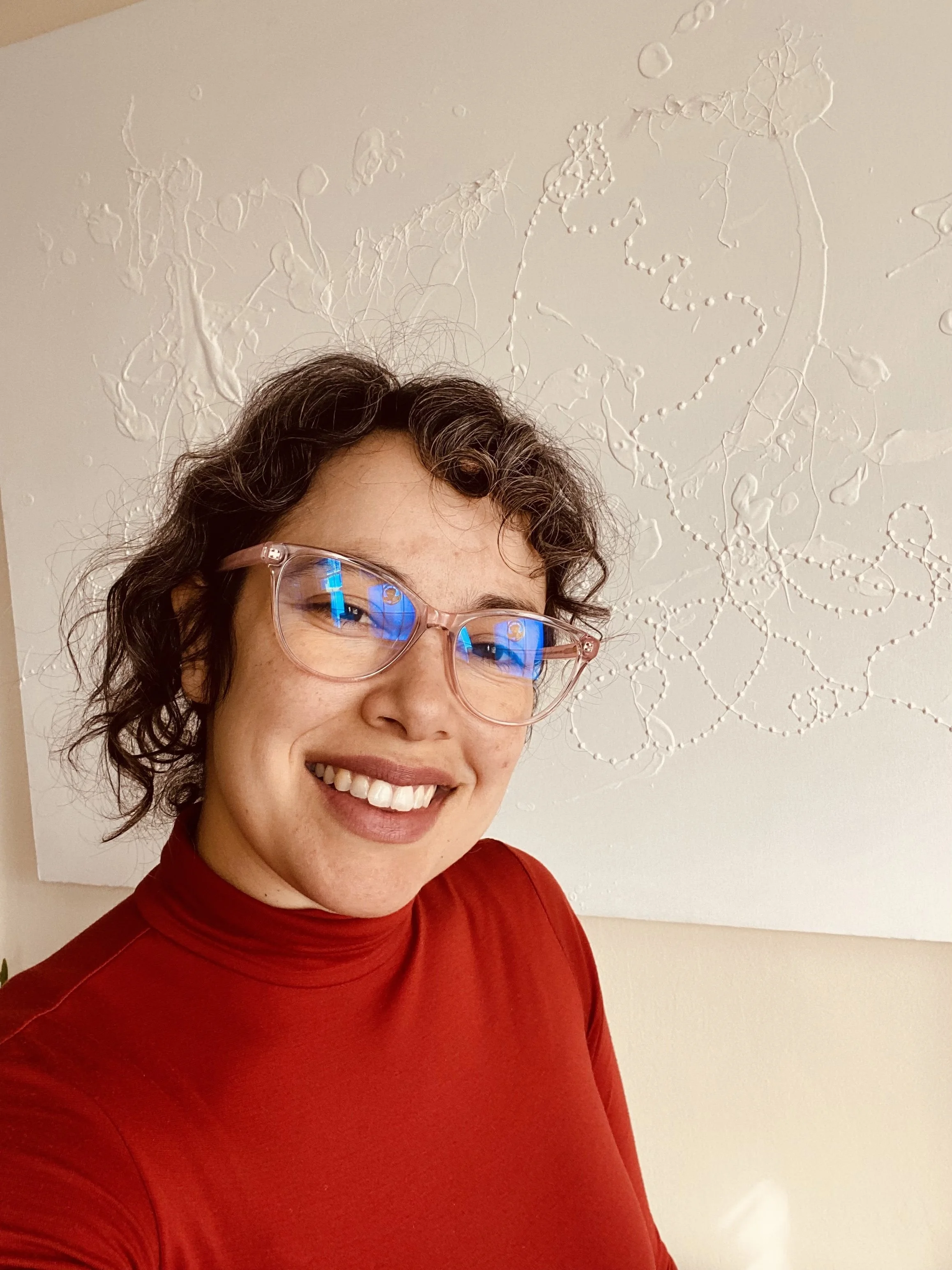 A woman with curly brown hair, wearing glasses with pink frames and a red turtleneck, smiling and standing in front of abstract white wall art.