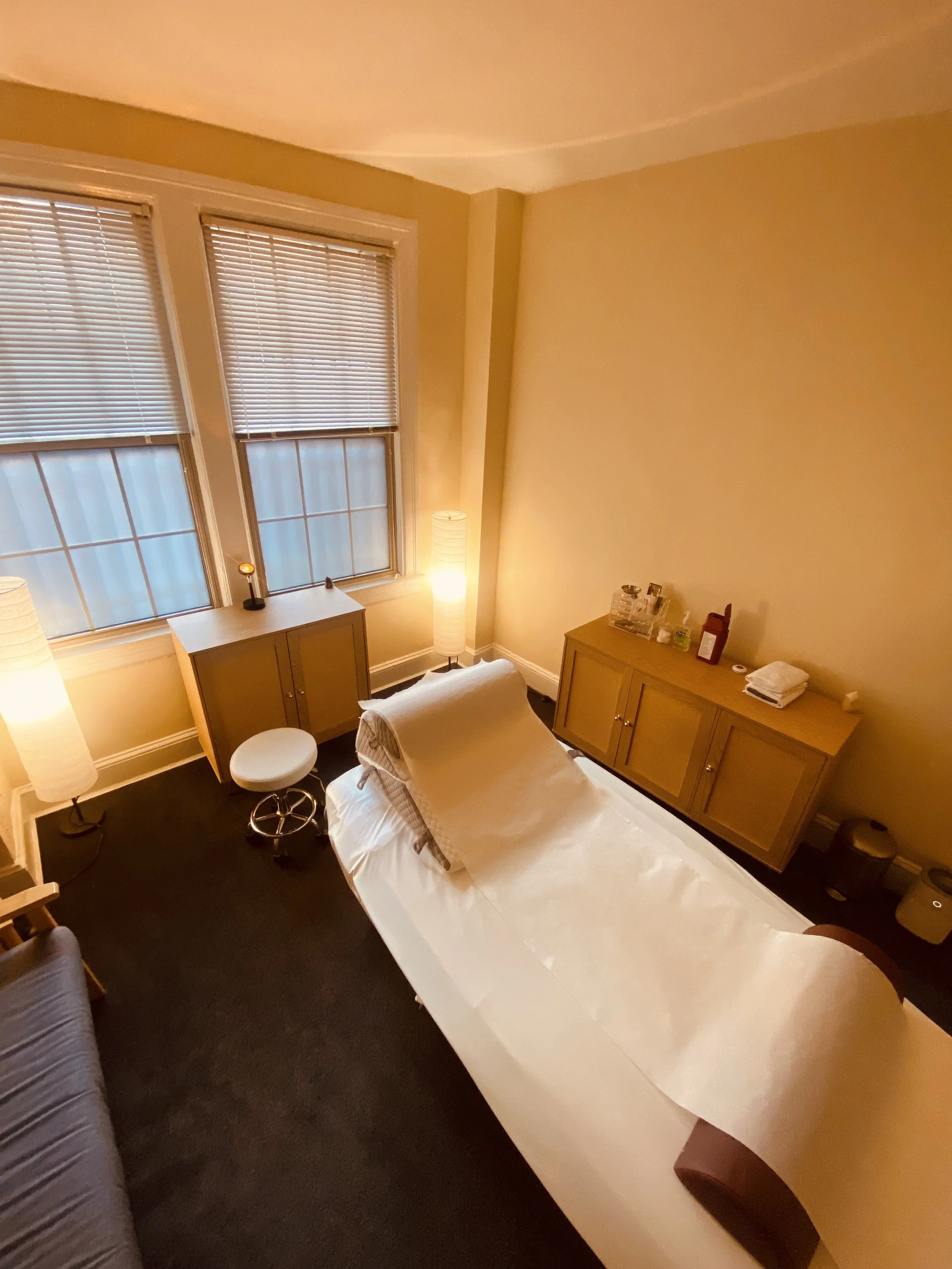 Image of a warm and inviting acupuncture treatment room, featuring a comfortable table with a supportive pillow, soft lighting, and frosted windows, as well as an organized supply table against the wall where the acupuncturist keeps their tools.