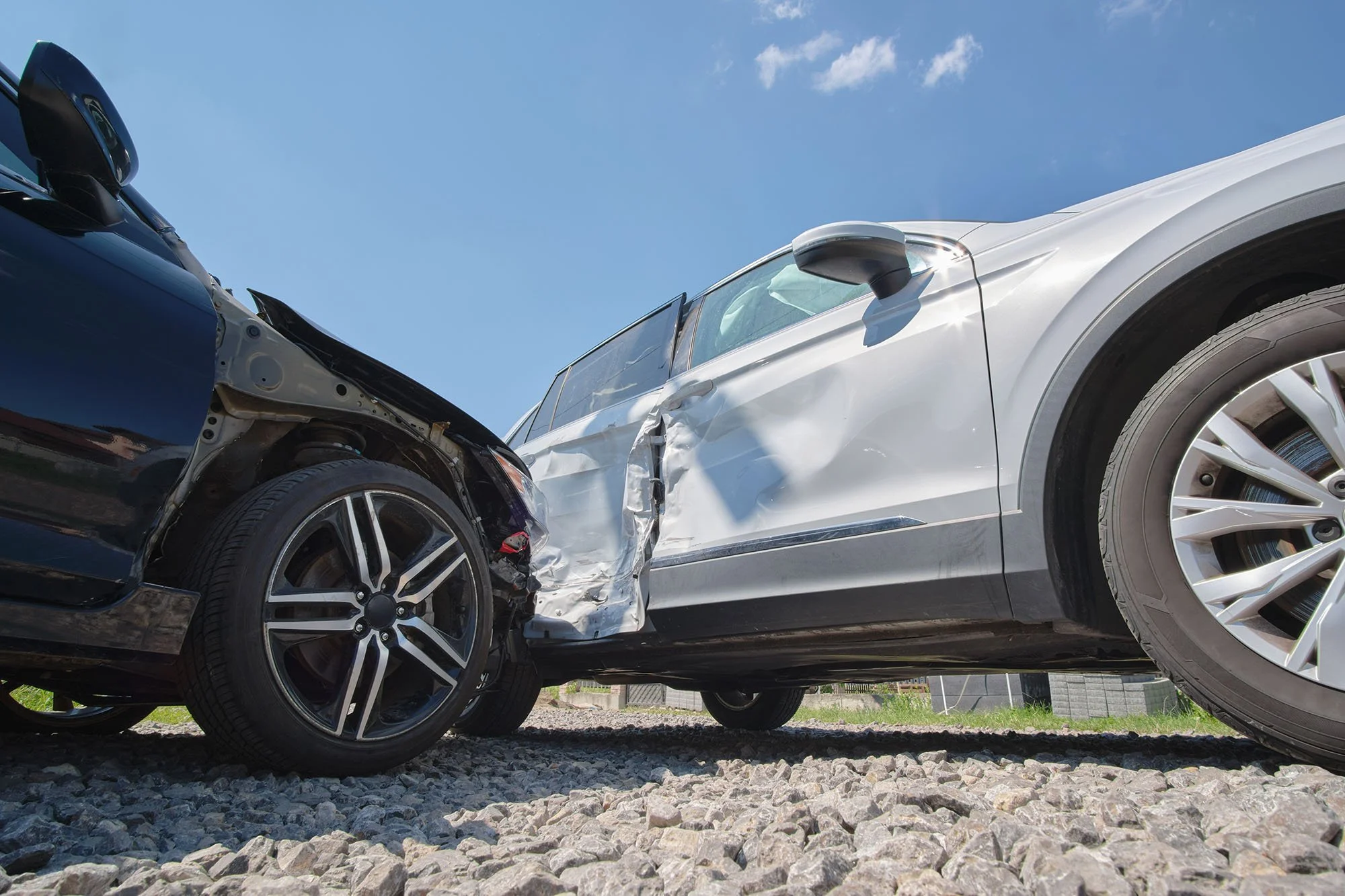 A black car and a silver car involved in a head-on collision with significant front-end damage on a gravel lot, under a blue sky with a few clouds.