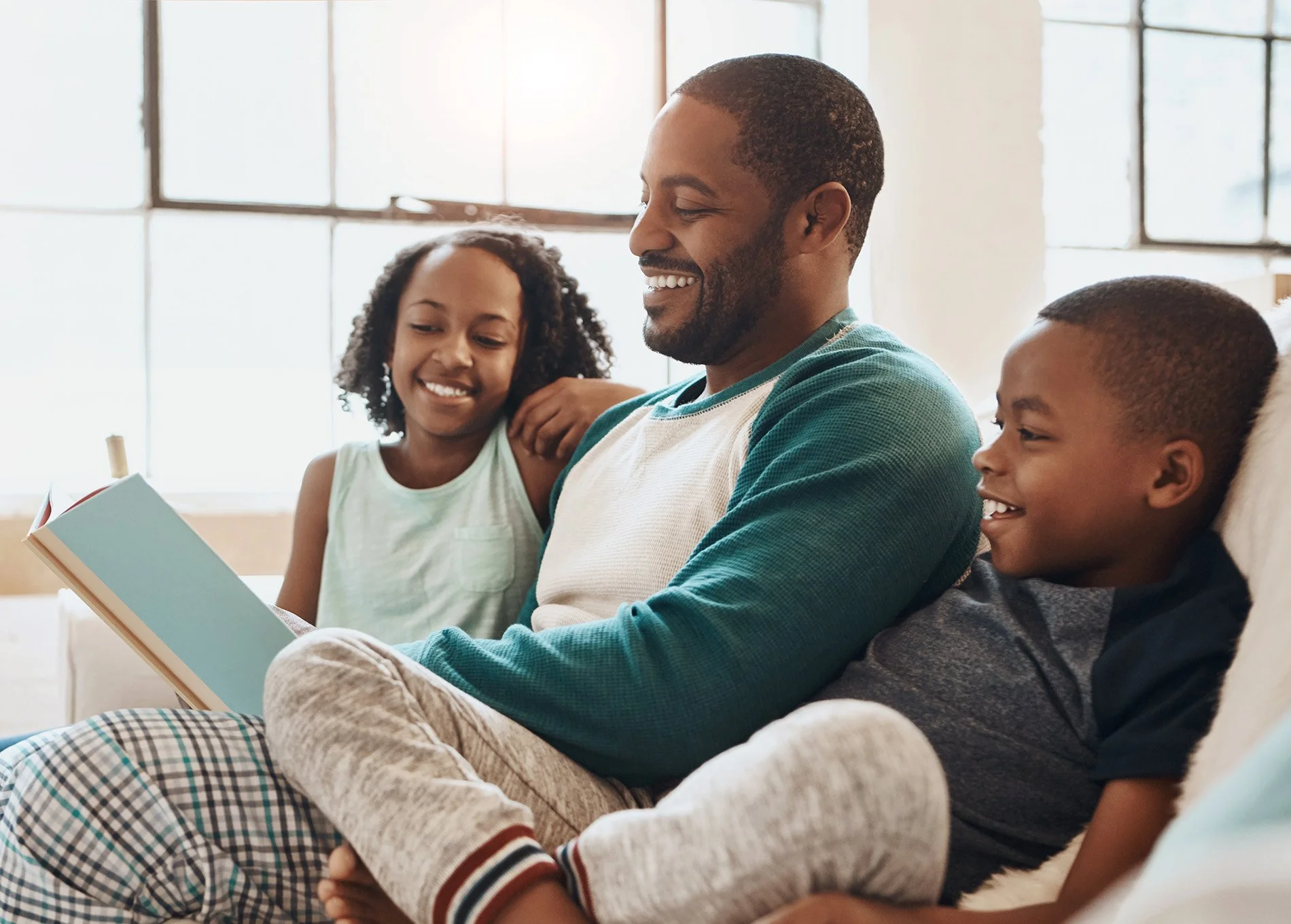 A man with two children sitting on a couch, reading a book together and smiling in a bright room with large windows.