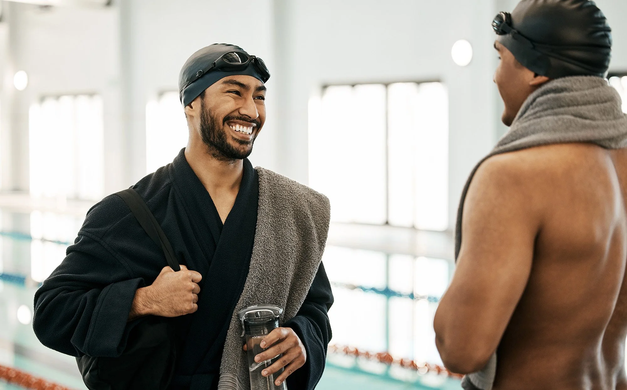 Two male swimmers wearing swim caps and goggles are engaging in conversation at an indoor swimming pool. One is smiling with a towel over his shoulder, holding a water bottle. The other is shirtless, facing away from the camera.