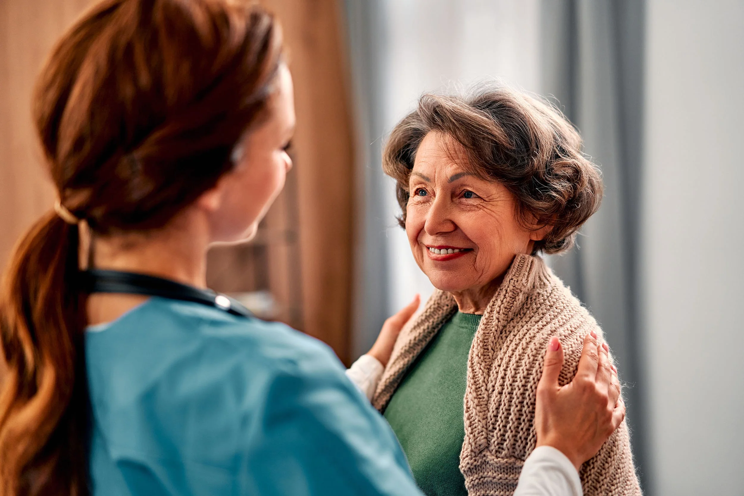 A healthcare worker smiling and comforting an elderly woman with gray hair, wearing a beige knitted sweater.