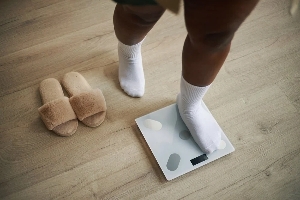 Person standing on a digital scale with white socks, next to beige fuzzy slippers on a wooden floor.
