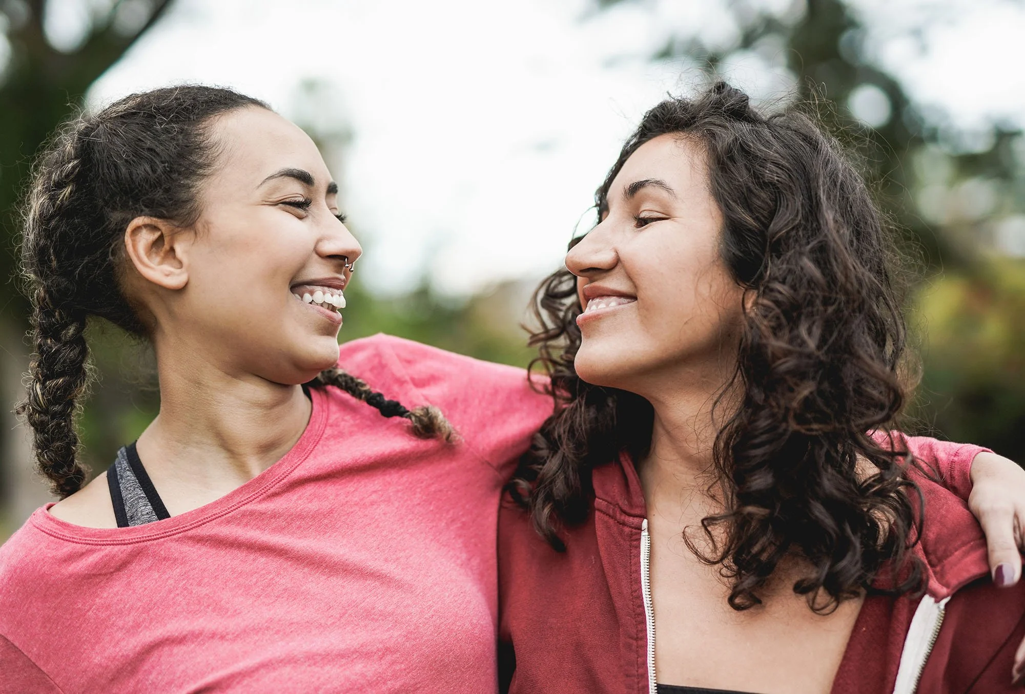 Two women smiling and embracing each other outdoors on a cloudy day, with blurred trees in the background.