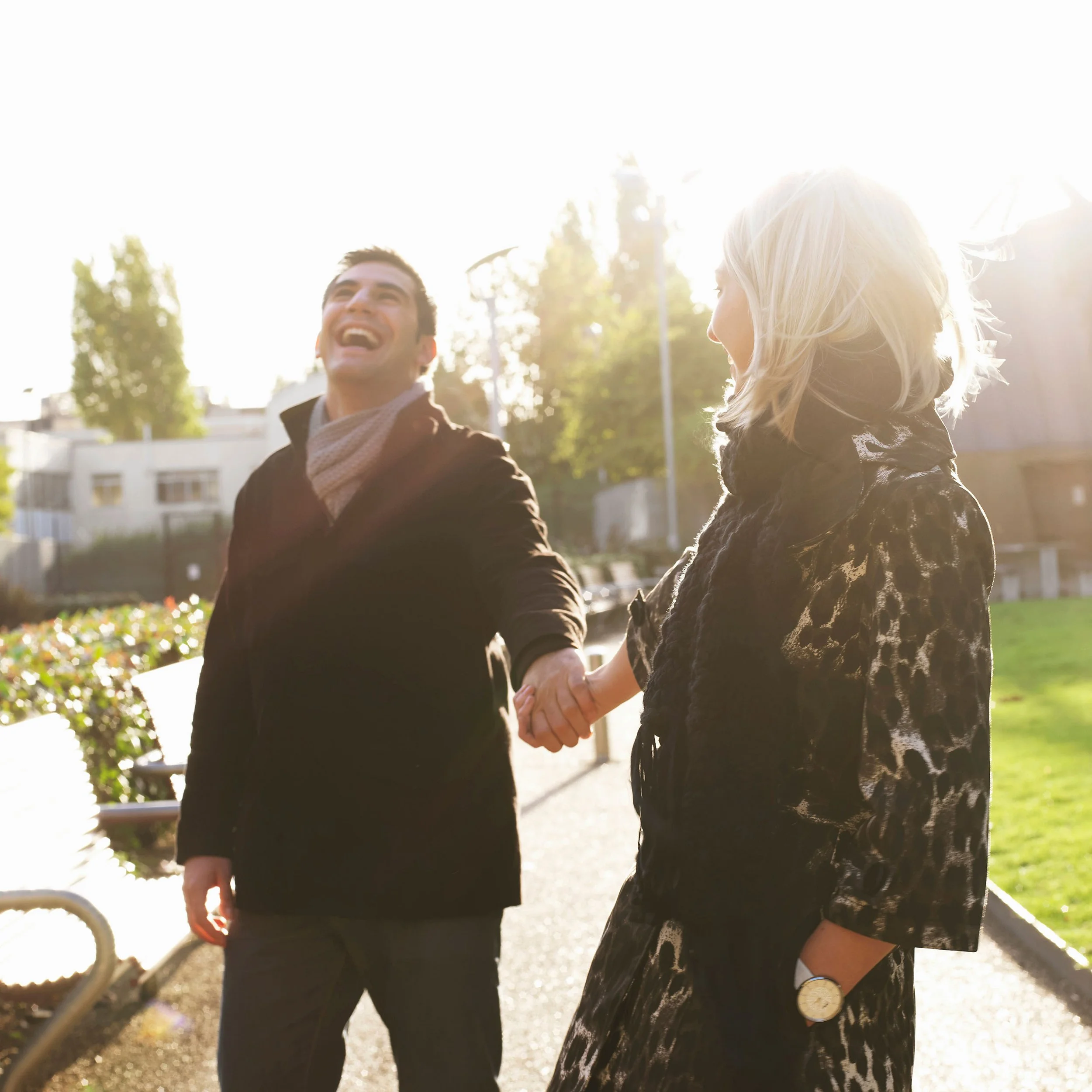Two people holding hands outdoors on a sunny day, with trees and a building in the background.