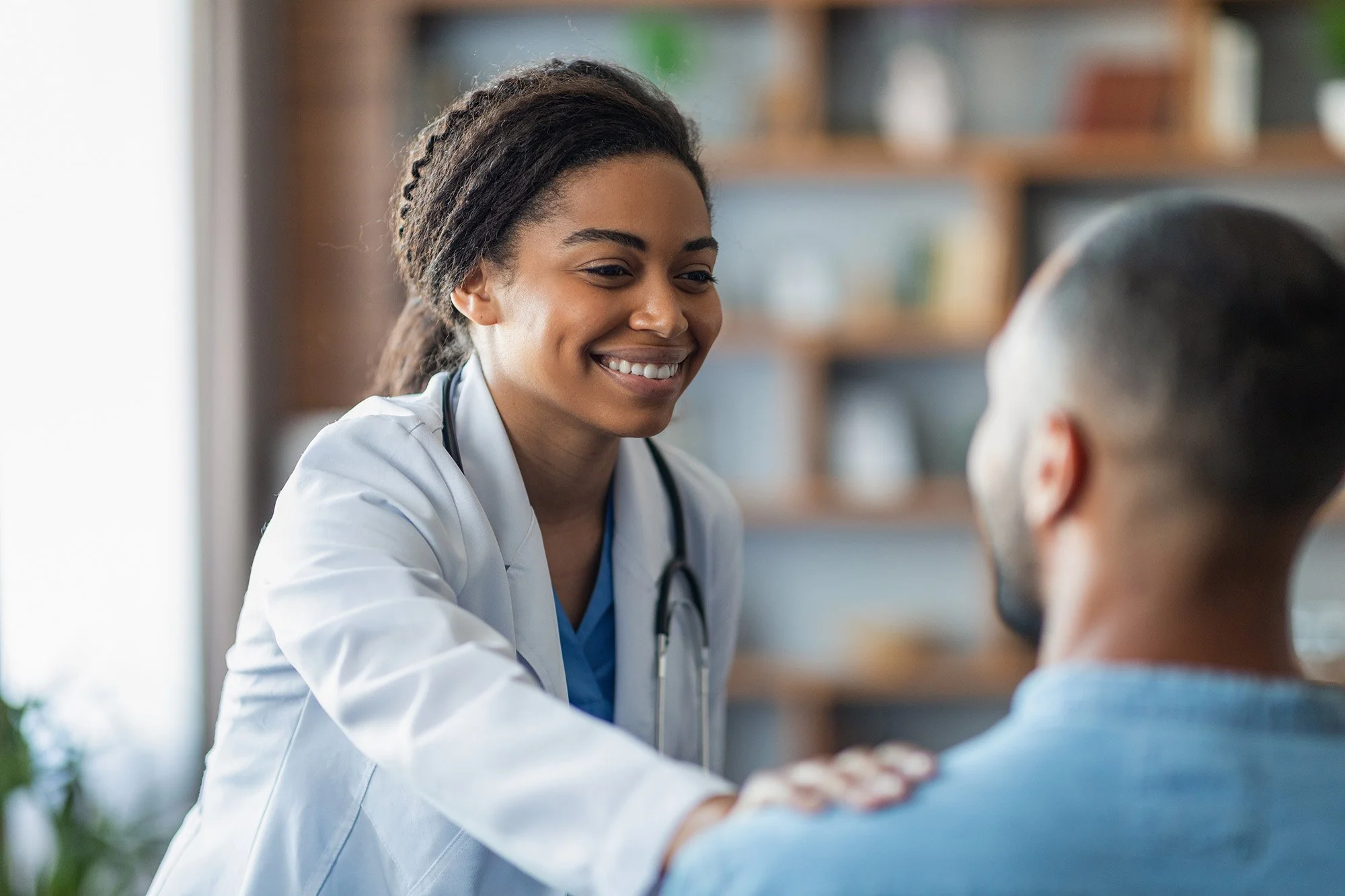A smiling female healthcare professional, wearing a white coat and stethoscope, engaging in a consultation with a patient in a clinic setting.