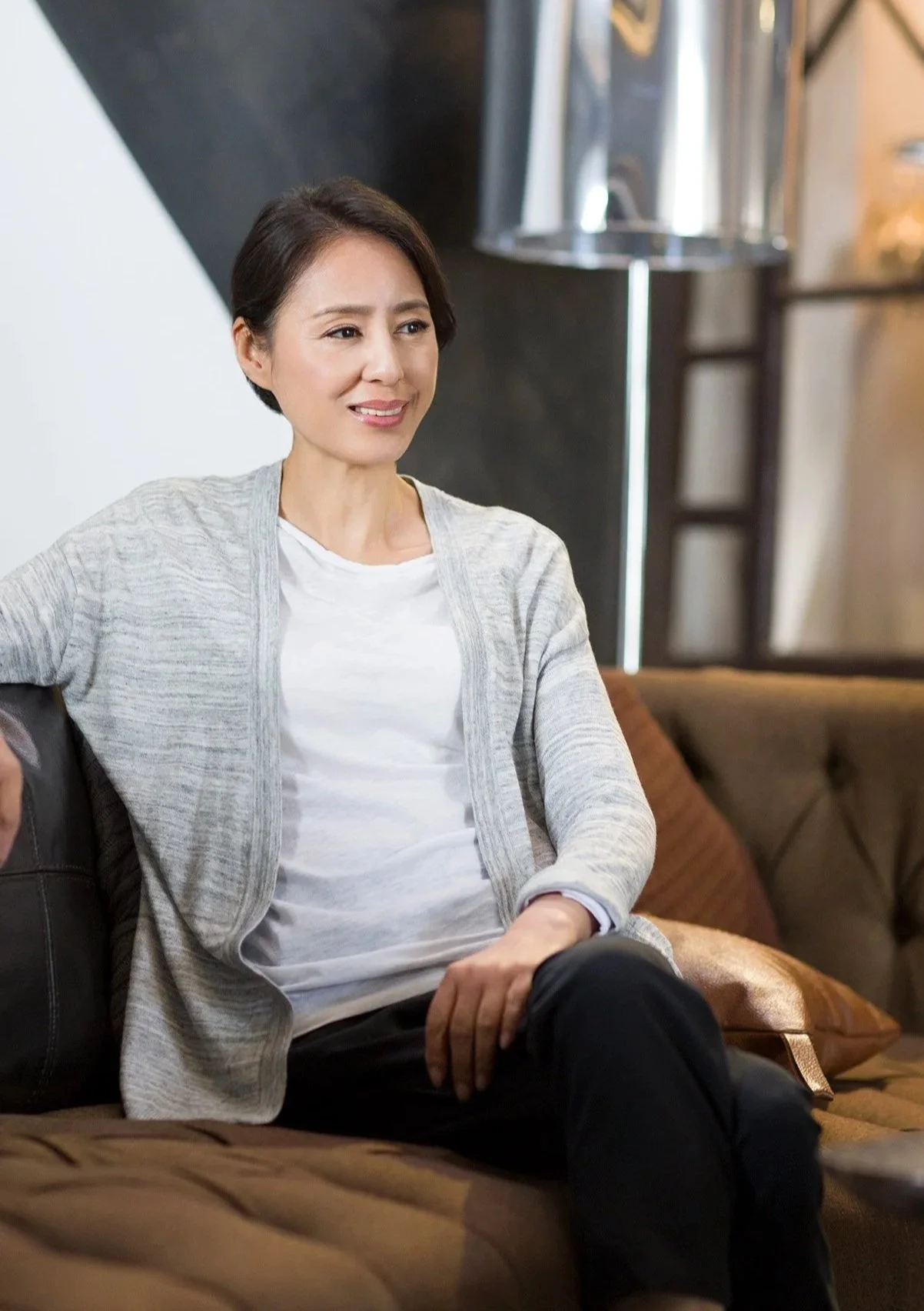 A woman with short dark hair sitting on a brown leather couch, smiling slightly, in a modern indoor space with a dark wall, a floor lamp, and decorative cushions.